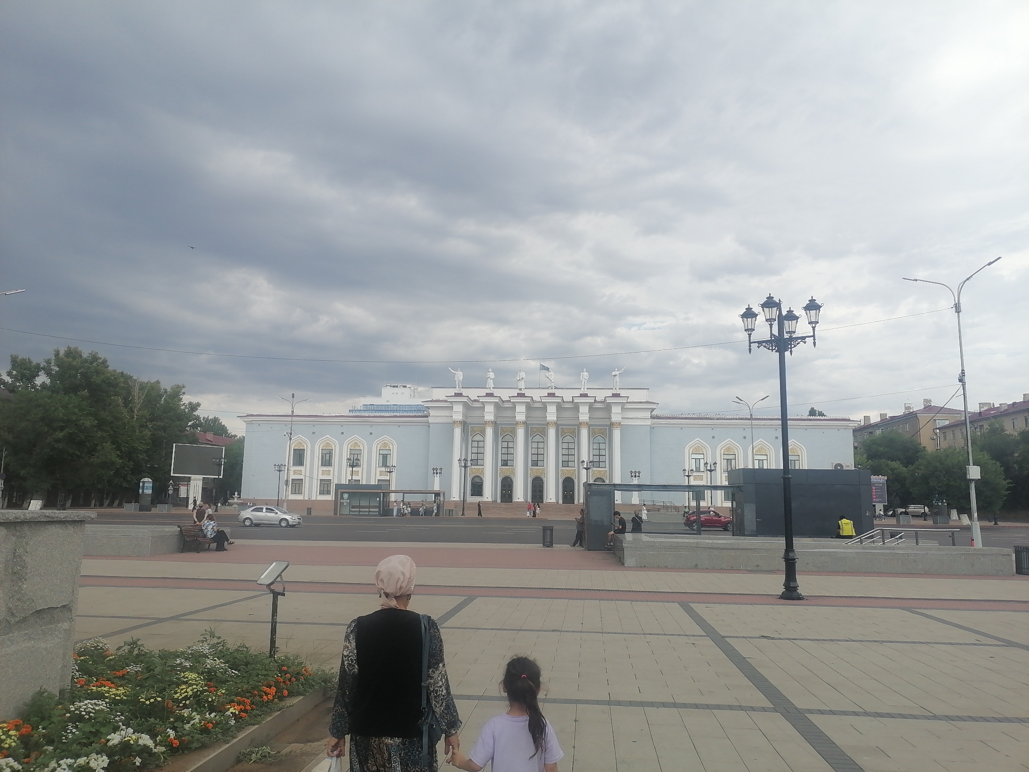 A cloudy day in Qarağandı, Kazakhstan, features the city administration building in the background, a grand structure with a light blue facade, white columns, and several statues on its roofline. In the foreground, a woman wearing a light pink headscarf and a patterned dress, and a young girl in a purple shirt are walking away from the camera. They are on a paved walkway in front of a flower bed with colorful blossoms. A street lamp with four lights stands to the right of the administration building. Several people are visible on benches near the building and on the street, where a silver car is also seen. A dark bus stop structure is situated on the sidewalk.