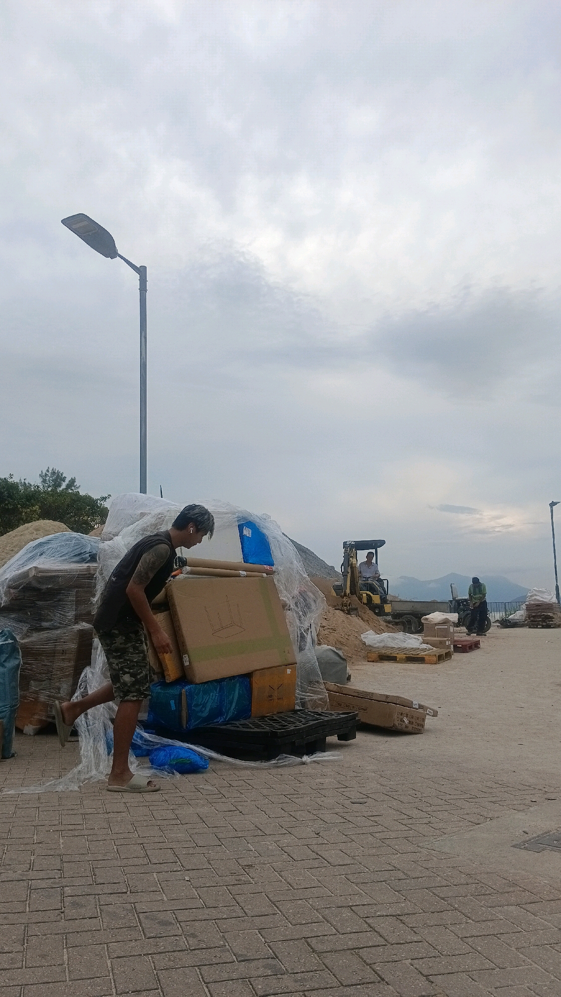 The image depicts an outdoor scene on a cloudy day, possibly in Hong Kong, China. In the foreground, a man wearing a black tank top and camouflage shorts is seen from the waist down, carrying a large cardboard box. He is wearing light-colored sandals. The man is standing on a paved pathway made of interlocking bricks. Behind him, there are large piles of materials, some covered in plastic sheeting, and several cardboard boxes and blue plastic-wrapped items are stacked.
In the mid-ground, a yellow excavator is visible, with a person sitting in the driver's seat. To the right of the excavator, another person is standing near a dark-colored vehicle. Further to the right, there are more stacks of materials and wooden pallets. The background shows a hazy mountainous landscape and the sea under a grey, overcast sky. A tall lamppost is prominent in the left mid-ground. The overall impression is of a construction or logistics site.