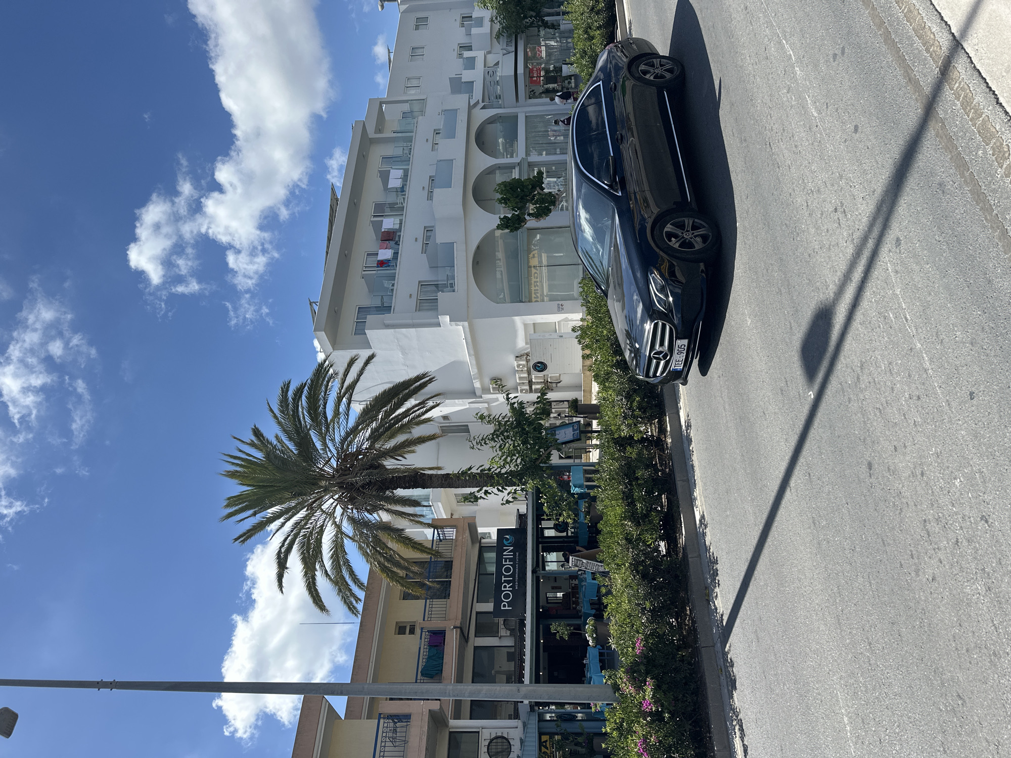 A black Mercedes-Benz E-Class sedan is driving on a paved road. The car has a license plate that reads "TEE 905". In the background, there is a street with a restaurant called "PORTOFINO" which serves "MEDITERRANEAN CUISINE". The restaurant has tables set with blue tablecloths outside. Behind the restaurant is a white multi-story building with balconies. A tall palm tree stands in front of the building. The sky is blue with some white clouds. The scene appears to be in a Mediterranean coastal town, likely in Paphos Municipality, Cyprus, given the style of architecture and vegetation. The overall impression is a bright, sunny day.