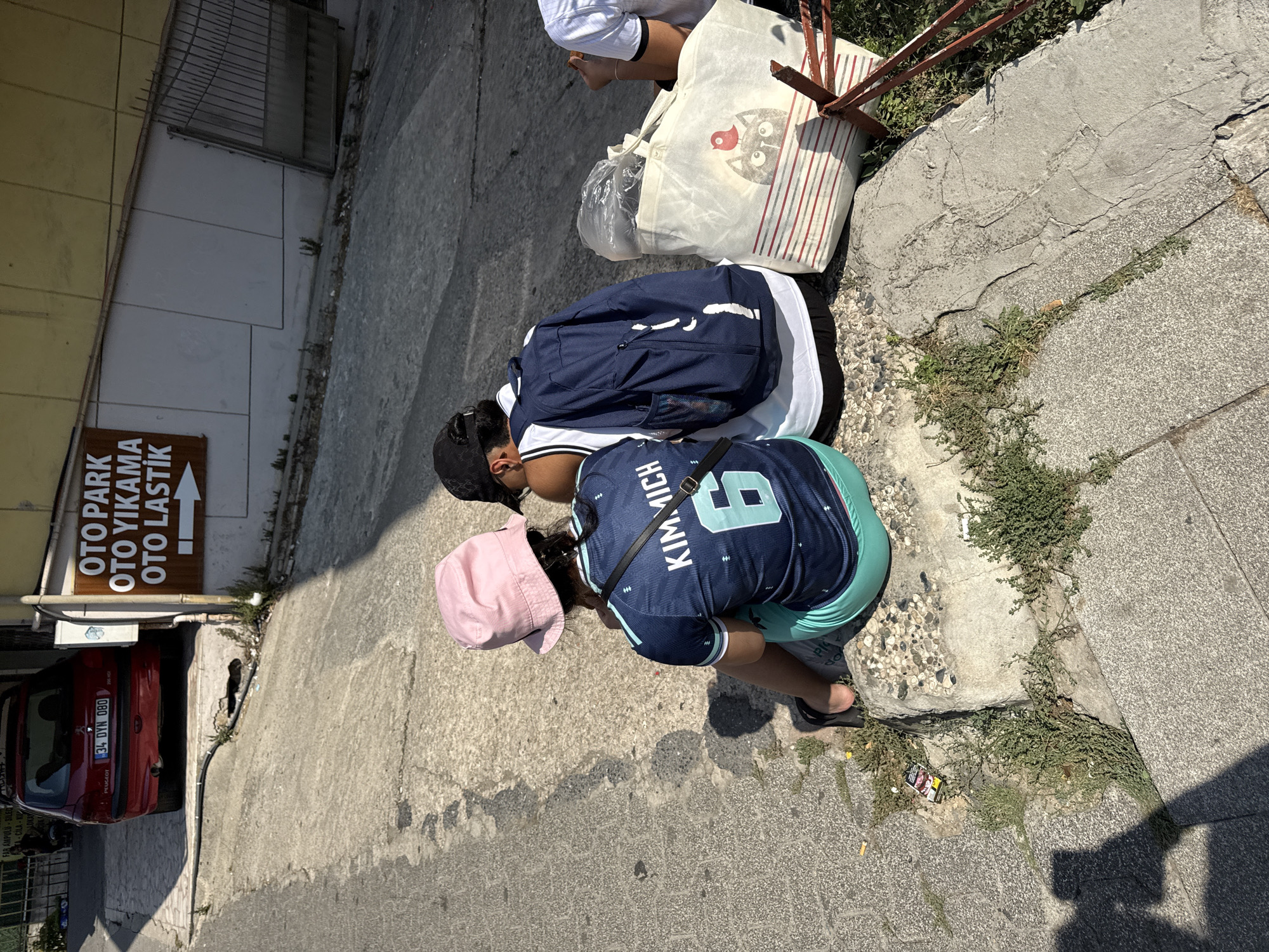 Two individuals, viewed from behind, are seated on a concrete ledge outdoors. The person on the left wears a pink bucket hat and a dark blue t-shirt with white lettering "KIM.MICHI" and the number "6" in light blue. They also wear light blue shorts. The person on the right wears a black baseball cap, a white tank top with black trim, and black pants. A dark blue backpack rests behind them. A white canvas tote bag with a graphic of a cat wearing a red hat and red and white striped accents is placed next to the tote bag. The setting appears to be a street in Kadıköy, Turkey, indicated by a sign in the background that reads "OTO PARK OTO YIKAMA OTO LASTİK" (Parking Lot, Car Wash, Car Tires) in Turkish. A red Peugeot car is parked behind the sign. The ground is paved with concrete and interspersed with patches of dry grass and weeds. A cigarette pack and several cigarette butts are visible on the ground near the seated individuals. The lighting suggests it is daytime.