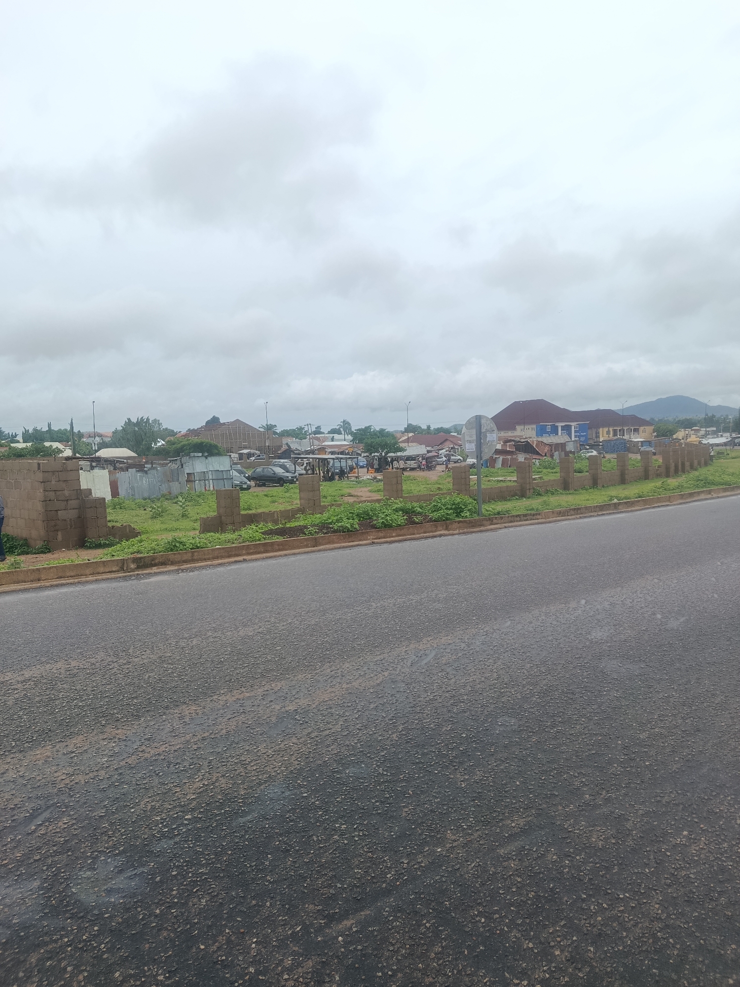 The image captures a street view in Jalingo, Nigeria, under an overcast sky. In the foreground, a paved road stretches across the frame, showing some wear and tear. To the left and slightly behind the road, a low, unfinished brick wall delineates a grassy area. Beyond this, a cluster of buildings, some appearing to be constructed with corrugated metal and others with more permanent materials, are visible. Several vehicles, including cars and possibly some vans, are parked or in transit among these structures. In the middle ground, more buildings rise, some with colorful facades, and a large, multi-story structure with a dark, sloping roof dominates the background. The landscape beyond is characterized by scattered trees and rolling hills, indicating a semi-urban or suburban setting on the outskirts of Jalingo. The overall impression is one of a developing area with a mix of residential and commercial structures, typical of many Nigerian cities.
