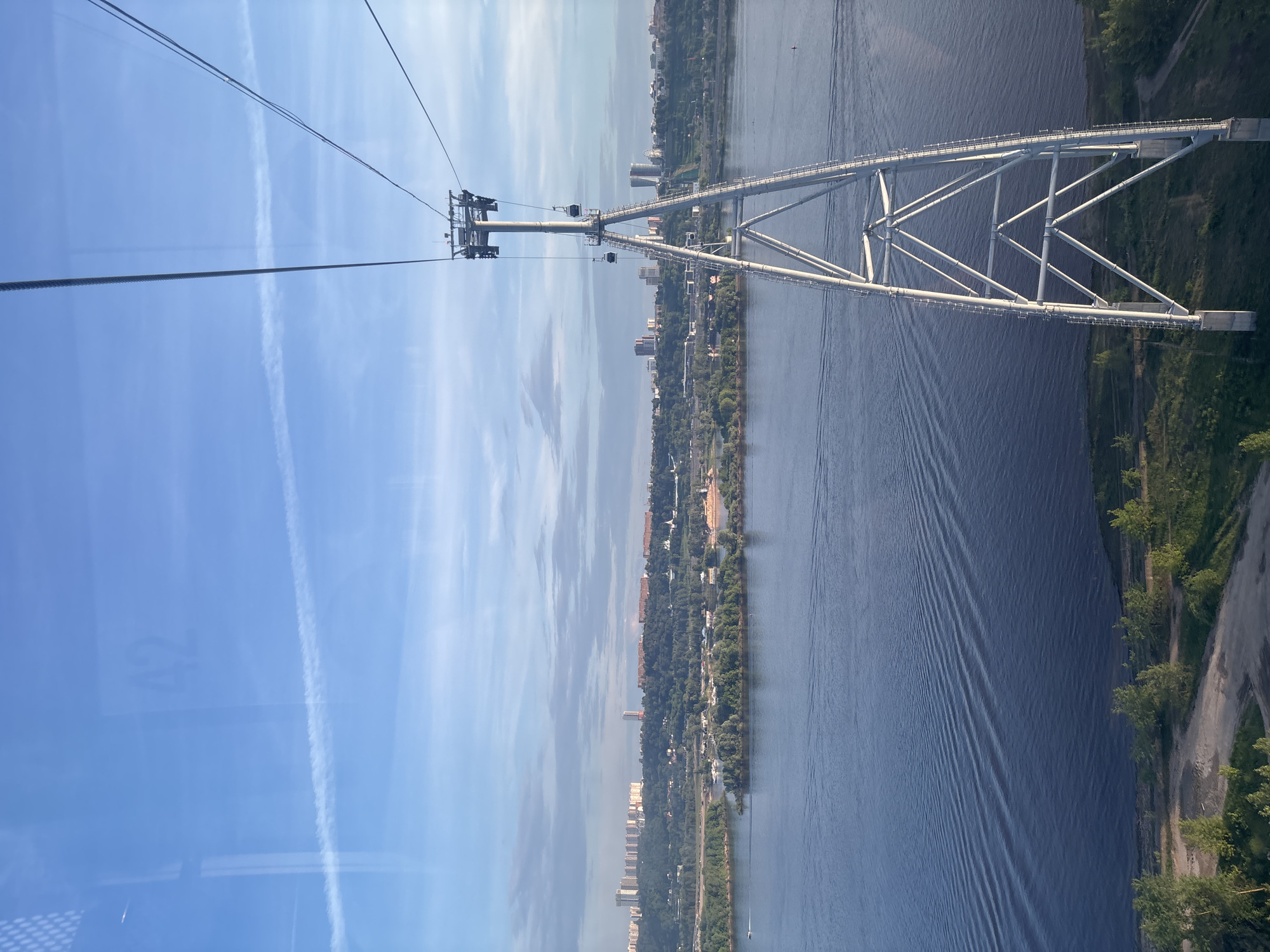 This image presents a panoramic view from a cable car, overlooking a broad river and its surrounding landscape. The perspective is elevated, characteristic of a ride high above the ground.
**Subjects:** The primary subjects include a vast river, a distinct white bridge, and elements of a cable car system. The river, likely the Volga, stretches across the middle ground, flanked by green forested banks and distant cityscapes with buildings. The bridge features a prominent white, open-truss structure, supported by multiple diagonal and horizontal beams. A tall cable car tower, part of the suspension system, stands on the left, with thick cables extending diagonally across the upper left portion of the frame. The sky is a prominent feature, occupying the upper half of the image.
**Activity or event taking place:** The image captures the experience of traveling by cable car, offering a scenic aerial view of the river and the urban-natural interface. Small ripples or wakes on the river surface suggest the presence of boats, though none are clearly discernible.
**Notable details:** The scene unfolds under a bright, clear blue sky with scattered wispy clouds and distinct white contrails, indicating a sunny daytime setting. The river's surface shows subtle patterns, possibly from water currents or passing vessels. Reflections of the sky and light are visible on the cable car window in the bottom left corner. Faintly visible on the window is the number "17", likely a cabin identifier. The overall impression is one of a vast, tranquil landscape viewed from a unique vantage point. Given the context, this is the Bor cable car system, providing a link across the Volga River near Bor, Russia.
**Visible text:** The number "17" is faintly discernible on the cable car's window surface.