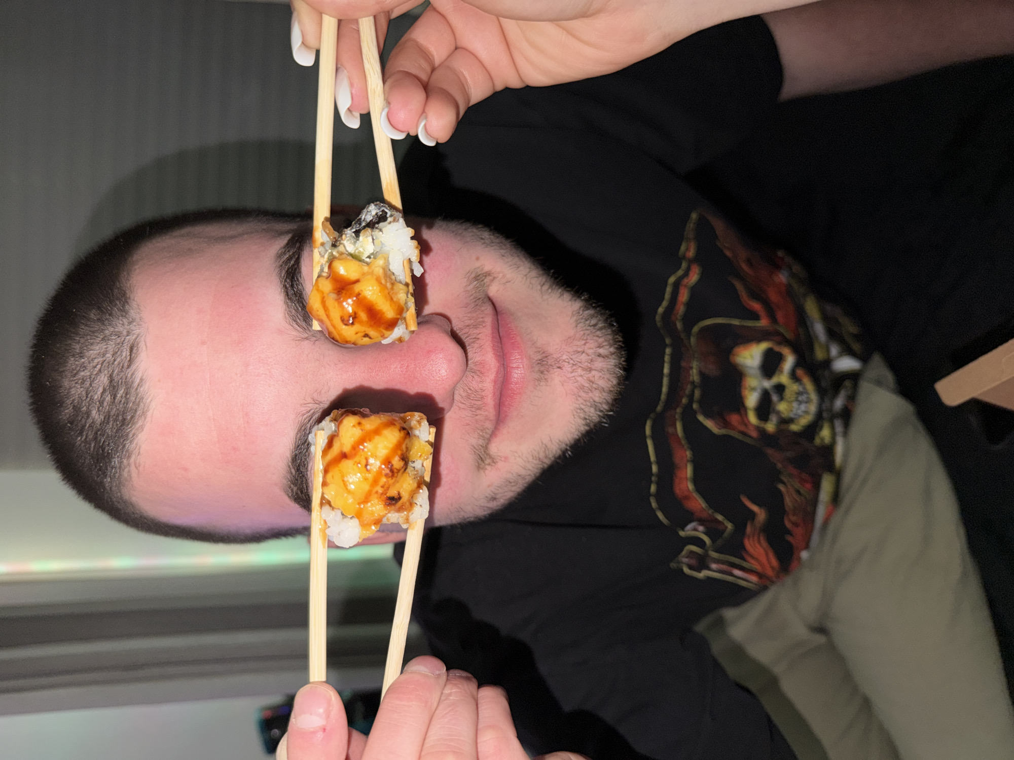 A man is holding two pieces of sushi with chopsticks in front of his eyes. He is wearing a black t-shirt with a skull design. The background is dark, with a glimpse of a light-colored item (possibly clothing) to the right. The location is not discernible from the image.
