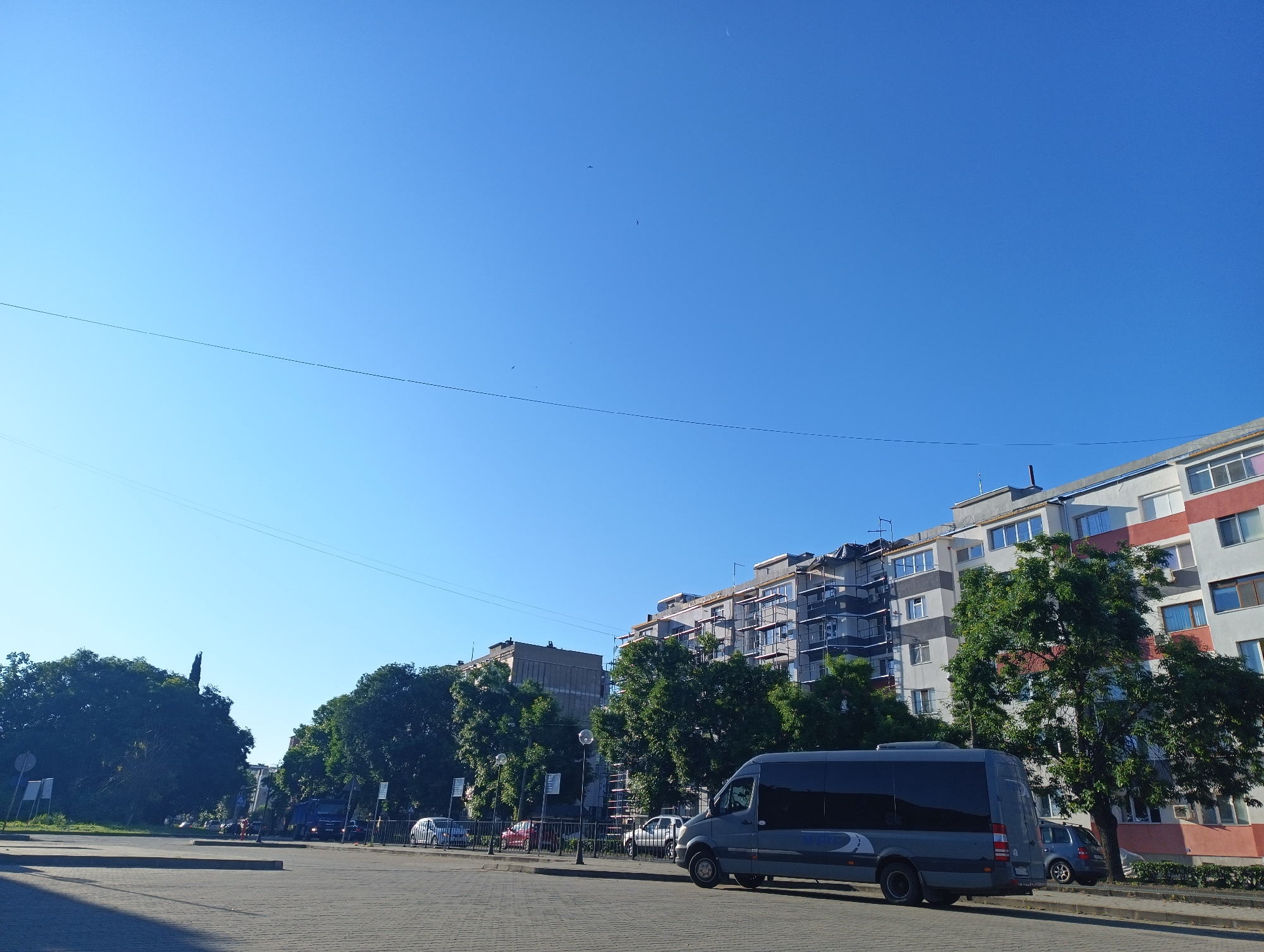 In Karnobat, Bulgaria, on a clear day, a gray passenger van is parked on a paved street. The van faces right, partially obscuring a row of parked cars. Beyond the vehicles, a tree-lined street leads to buildings with a mix of modern and older architectural styles. The buildings are several stories high and some appear to be under renovation, with scaffolding visible. The sky is a bright, cloudless blue.