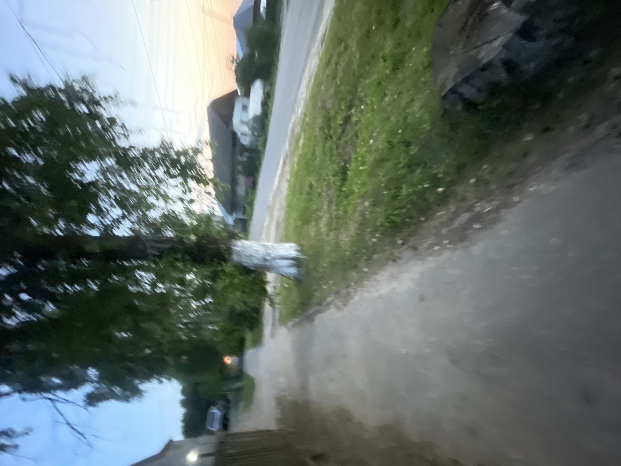 The image depicts a street scene in Krasnye Baki, Russia, likely during dusk or dawn, given the soft lighting and colorful sky. A paved sidewalk runs along the left side of the image, bordered by a grassy area on the right. A tree with a whitewashed trunk stands prominently on the right side of the sidewalk. Beyond the grassy area and sidewalk, a road is visible, with houses and other structures lining the opposite side. One house appears to have a dark roof and white siding, with a white trailer or camper parked in front of it. The sky transitions from a soft blue to hues of orange and pink near the horizon. The overall impression is that of a quiet, semi-rural residential area.