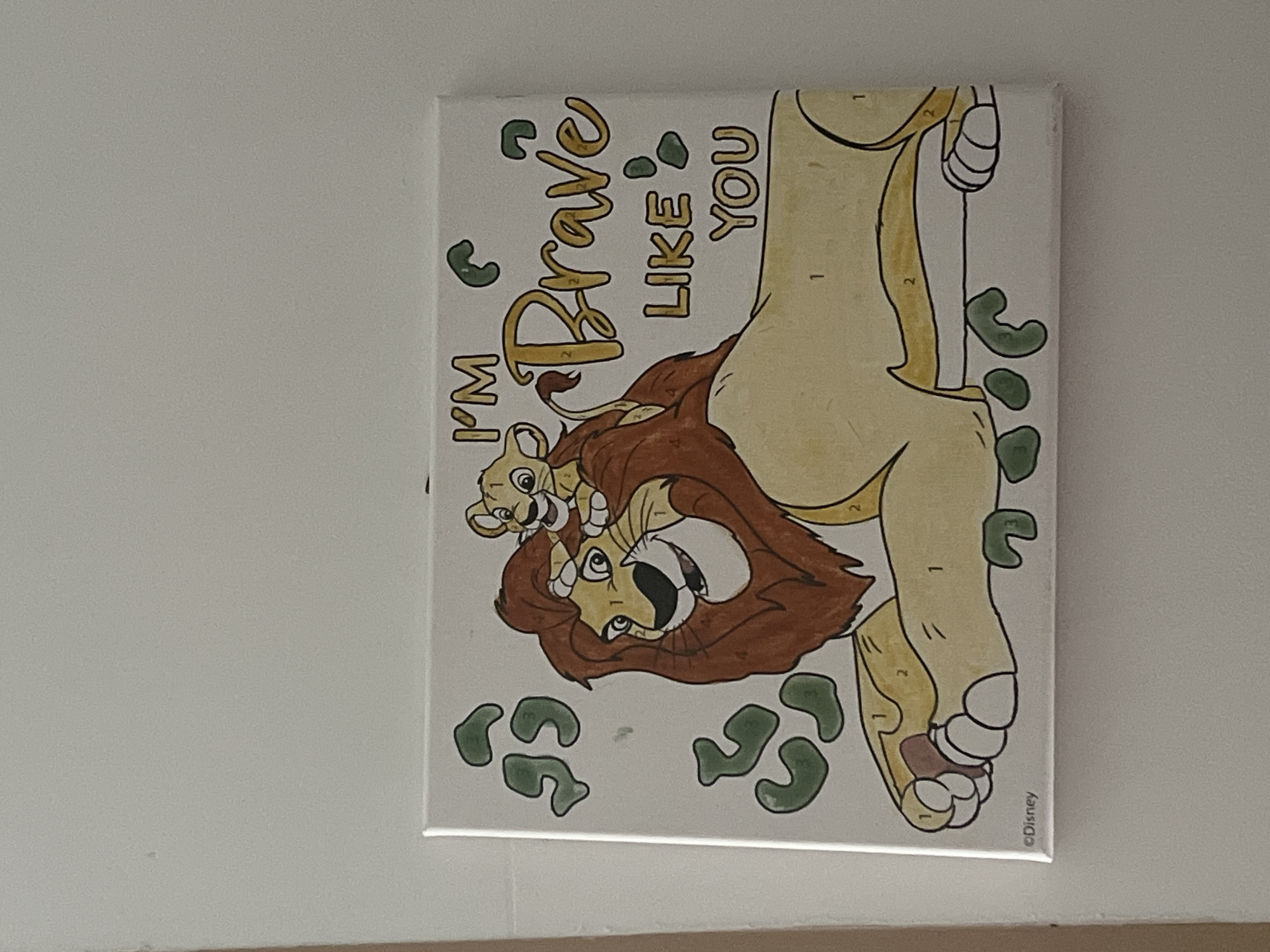 The image displays a paint-by-numbers canvas featuring characters from Disney's The Lion King. Simba, a young lion cub, is perched on the head of Mufasa, an adult lion. The text "I'M BRAVE LIKE YOU" is written in large, stylized letters on the canvas. Scattered green shapes, resembling leaves or paw prints, are visible around the main figures. The bottom left corner of the canvas bears the Disney copyright mark. The canvas is mounted on a light-colored wall. The background suggests an indoor environment, possibly a home, given the plain wall. There are no visible elements that would definitively place this scene within the city of Kohtla-Järve linn, Estonia. The lighting appears to be ambient indoor lighting.