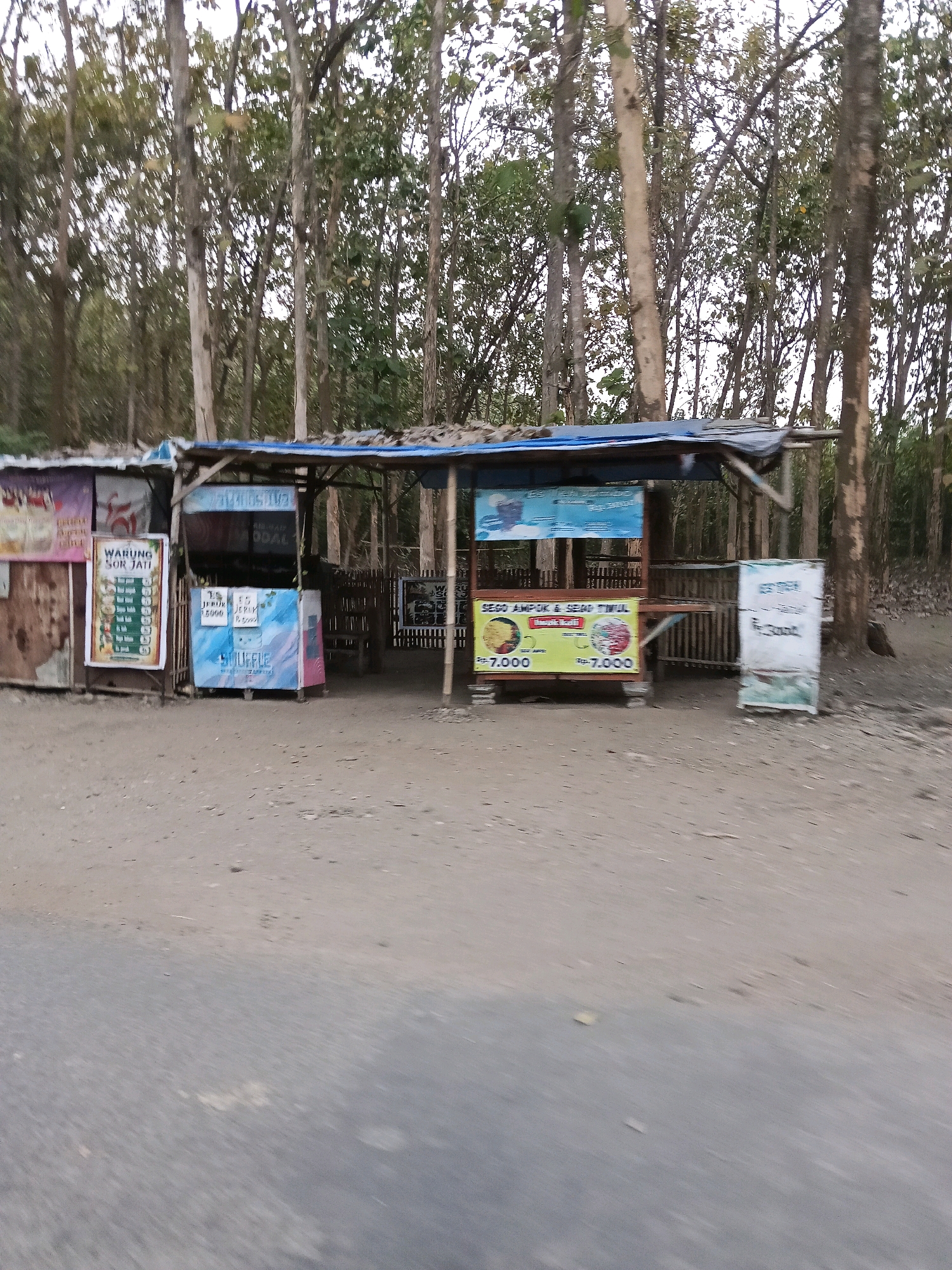 The image shows a roadside food stall in Gogodeso, Indonesia. The stall is constructed from wood and covered with a blue tarp. It is situated in front of a dense forest of teak trees.
There are several signs displayed at the stall. One sign, on the left, is pink and appears to be a menu or advertisement. Next to it is a vertical sign with the text "WARUNG SOR JATI" and a list of items with prices. In the center, there are two yellow signs, each displaying a picture of food and priced at "Rp. 7,000". Another sign, on the far right, is white with some text and graphics, but it is difficult to read clearly.
In front of the stall is a dirt and gravel area, with a paved road in the foreground. The lighting suggests it is daytime, possibly late afternoon, as the light is somewhat diffused. There are no people visible in the image. The overall scene depicts a simple, rustic food vendor typical of rural Indonesian settings.