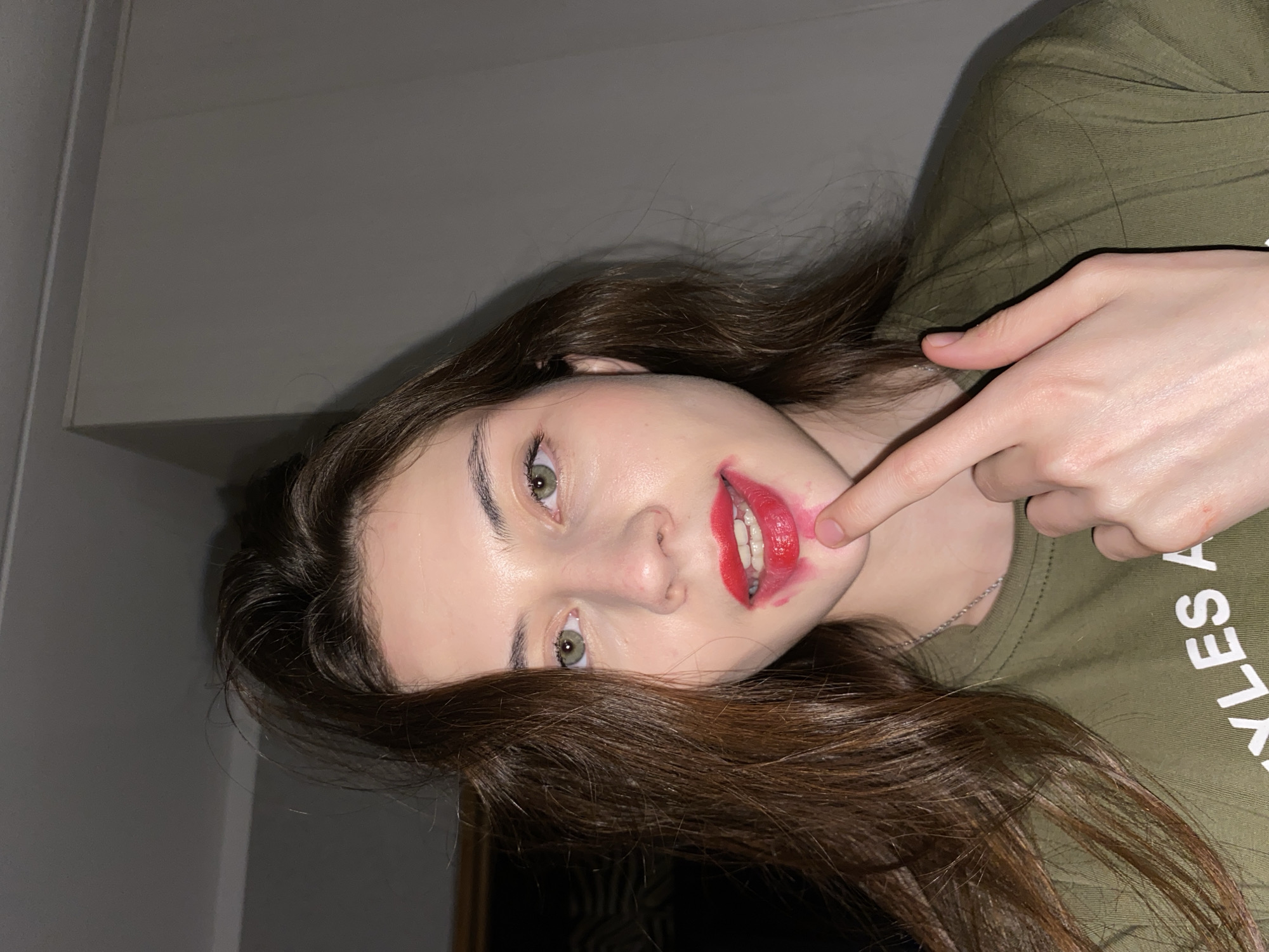 A young woman is depicted in a close-up, waist-up shot, looking directly at the camera with a neutral expression. Her head is tilted, suggesting she might be lying down or the image is rotated. She has long, dark brown hair, fair skin, and striking green eyes. A prominent feature is her bright red lipstick, which is messily smeared around her mouth, extending onto her chin and cheek.
She wears a dark olive-green t-shirt, and a thin silver chain necklace is visible around her neck. Her left index finger is pointing towards the smeared lipstick on her lower lip and chin, drawing attention to it. Partial white text is visible on her t-shirt, appearing to spell out "YLES A" or "STYLES A" and "LES".
The background is a plain, light grey or off-white wall, indicating an indoor setting. The lighting is soft and appears to be artificial, typical of an interior space. There are no specific cues for the time of day or weather. The overall mood conveyed by her direct gaze and the playful gesture towards her smudged lipstick suggests a self-aware, casual, or possibly mischievous moment. The scene takes place in Votkinsk, Russia.