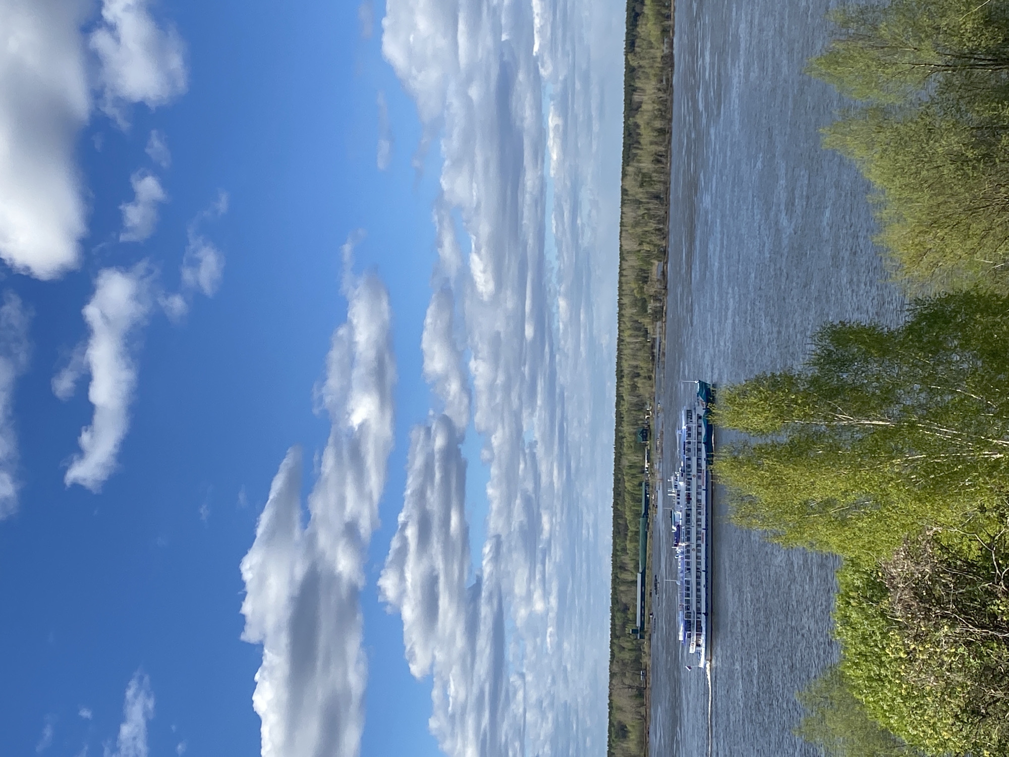 The image shows a river landscape in Ольховка, Russia. A passenger ship is docked on the right side of the river, near green trees and vegetation. On the opposite side of the river, a strip of dense forest runs along the bank. The sky is predominantly blue with scattered cumulus clouds.