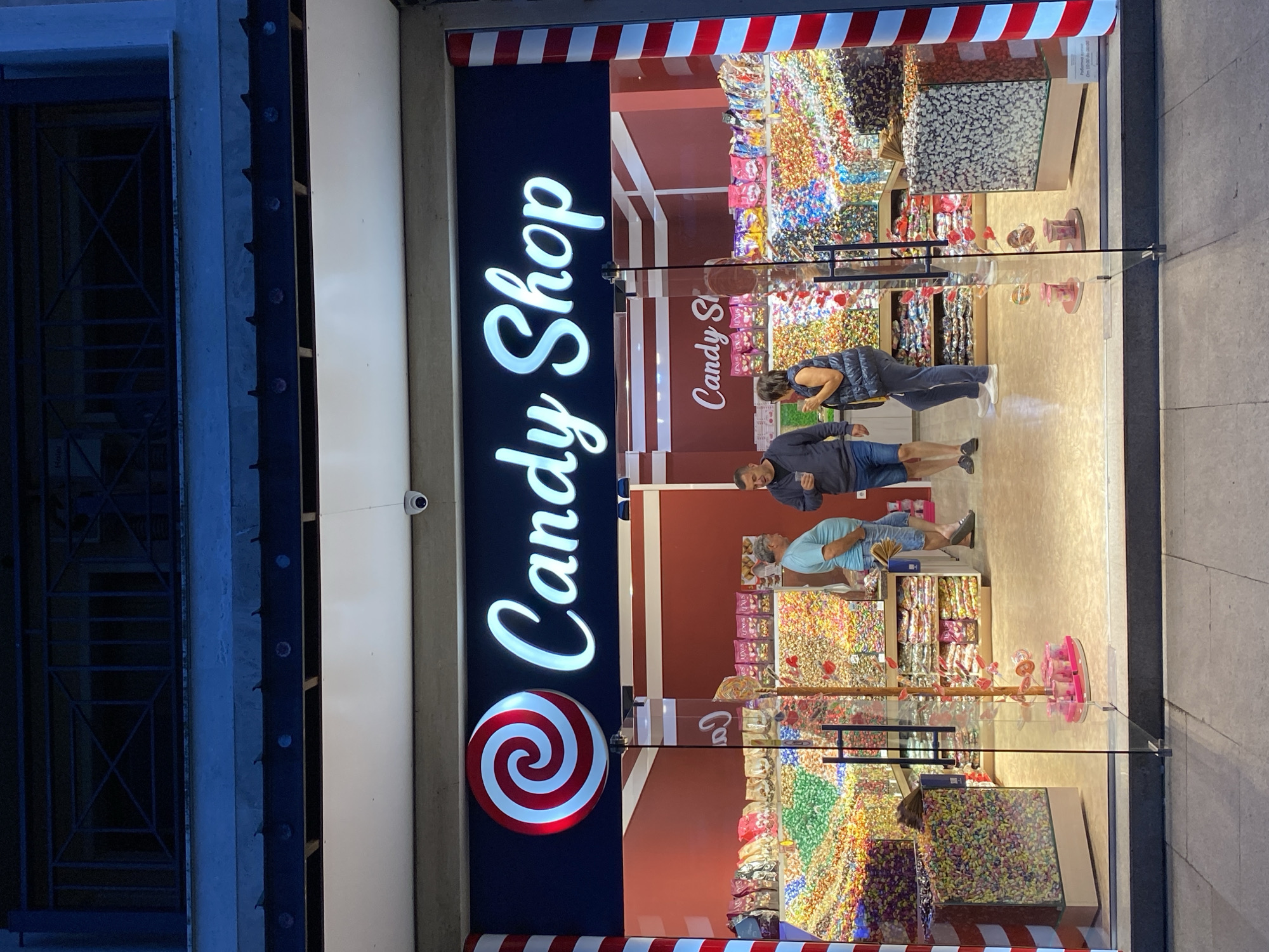 The image displays the brightly lit storefront of a "Candy Shop" at night in Varna, Bulgaria. The shop's name, "Candy Shop," is prominently displayed in white illuminated script on a dark blue fascia, accompanied by a large red and white spiral lollipop logo on the left. A white dome-shaped surveillance camera is mounted on the ceiling above the entrance. The shop entrance features glass double doors flanked by vertical columns adorned with red and white candy cane stripes. A partially visible white sign with Cyrillic text, confirming "Работно време / От 10:00 до 24:00" (Working hours / From 10:00 to 24:00), is positioned to the right inside the shop.
Three individuals are visible inside the shop, viewed through the glass. From left to right:
1. A male with short dark hair, wearing a dark top and shorts, holding a clear cup in his left hand.
2. A male with grey hair, wearing a light blue polo shirt and light blue denim shorts, with his right hand in his pocket. He is facing right, appearing to interact with the third person.
3. A female with dark hair, wearing a dark puffy vest over a lighter top and dark pants, holding a dark item in her left hand. She is facing left, appearing to interact with the middle male.
The shop's interior walls are reddish-brown. Extensive displays of bulk candies are visible in clear bins and shelves along the back and sides, featuring various colors and types. Packaged candies, some bearing the partial brand name "LEXUS," are arranged on shelves above the bulk displays. Two large, clear, rectangular bins filled with small, colorful candies are positioned near the front corners of the shop. In the center, a decorative pole-like structure with small candy items on pink stands is visible between the double doors. Above the shopfront, a dark architectural element with decorative projections and a dark building facade with a metal balcony railing are present.