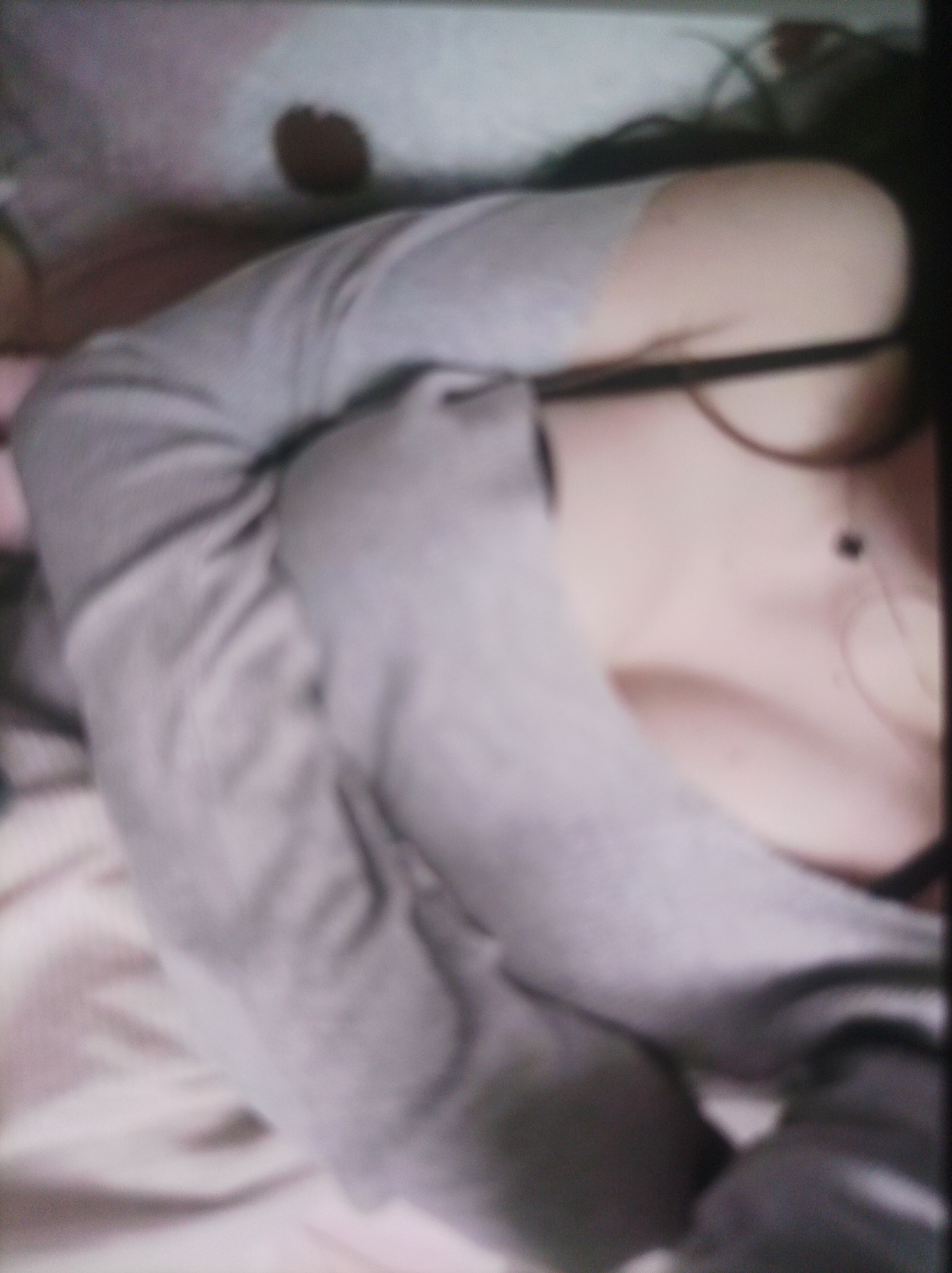 The image is a close-up, blurry shot of a person lying down, wearing a grey long-sleeved shirt. The person's torso and arm are visible, and they appear to be on a bed or soft surface with light-colored bedding. The lighting is dim and the focus is soft, obscuring fine details. There are no discernible elements that would indicate a specific location in a city.