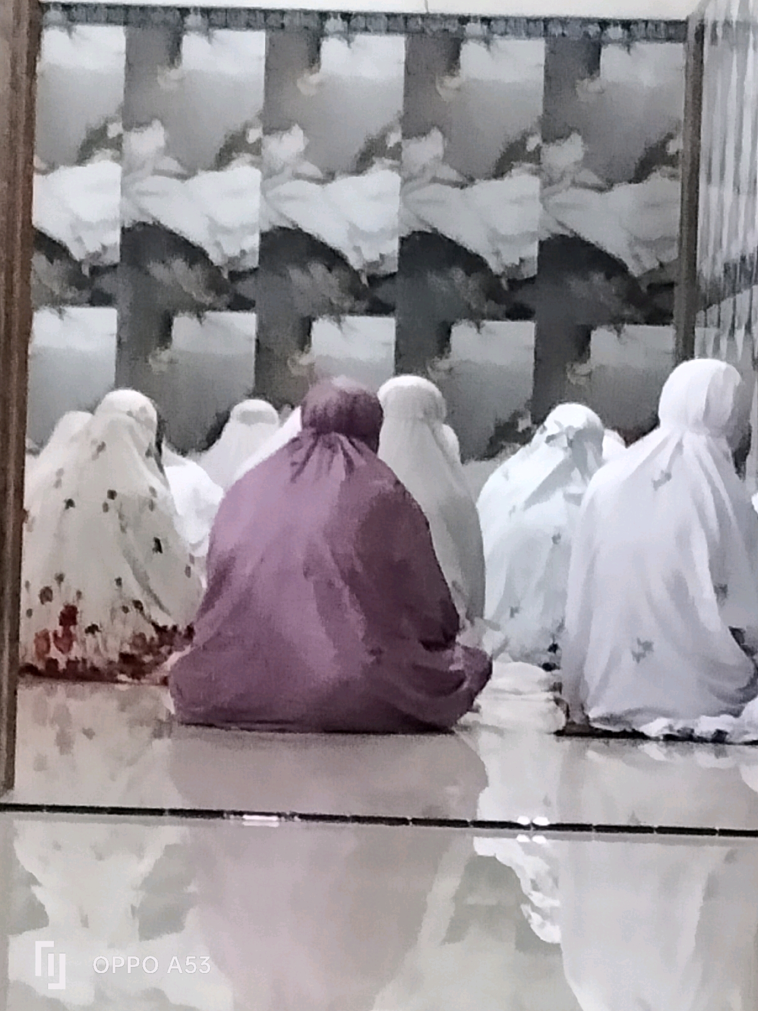 A group of individuals, all wearing prayer garments that cover their bodies and heads, are seated on a shiny tiled floor. The prayer garments vary in color, with one being a deep purple, another white with a floral pattern, and others plain white. The individuals are facing away from the camera, suggesting they are engaged in prayer. The background features a patterned wallpaper with an abstract, possibly floral or cloud-like design. The lighting is dim, creating reflections on the floor. The scene appears to be indoors, possibly in a prayer room or a home.