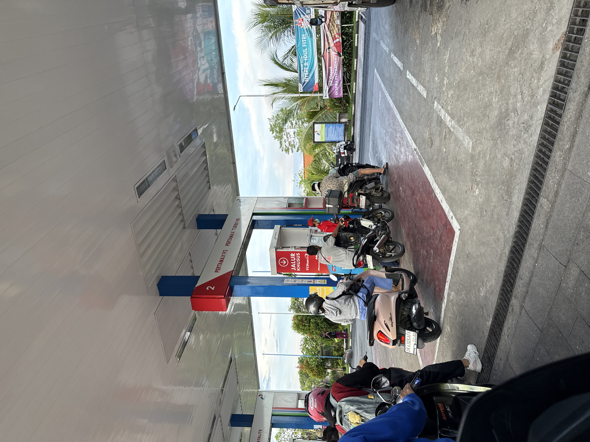 The image shows a gas station in Ungasan, Indonesia. Several people on motorbikes are queued at the gas pumps. The pumps are branded with the "Pertamina" logo. Banners wishing "Selamat Hari Raya Nyepi & Idul Fitri" are visible in the background. Pump number 2 provides Pertamax 92 and Pertamax Turbo 98.