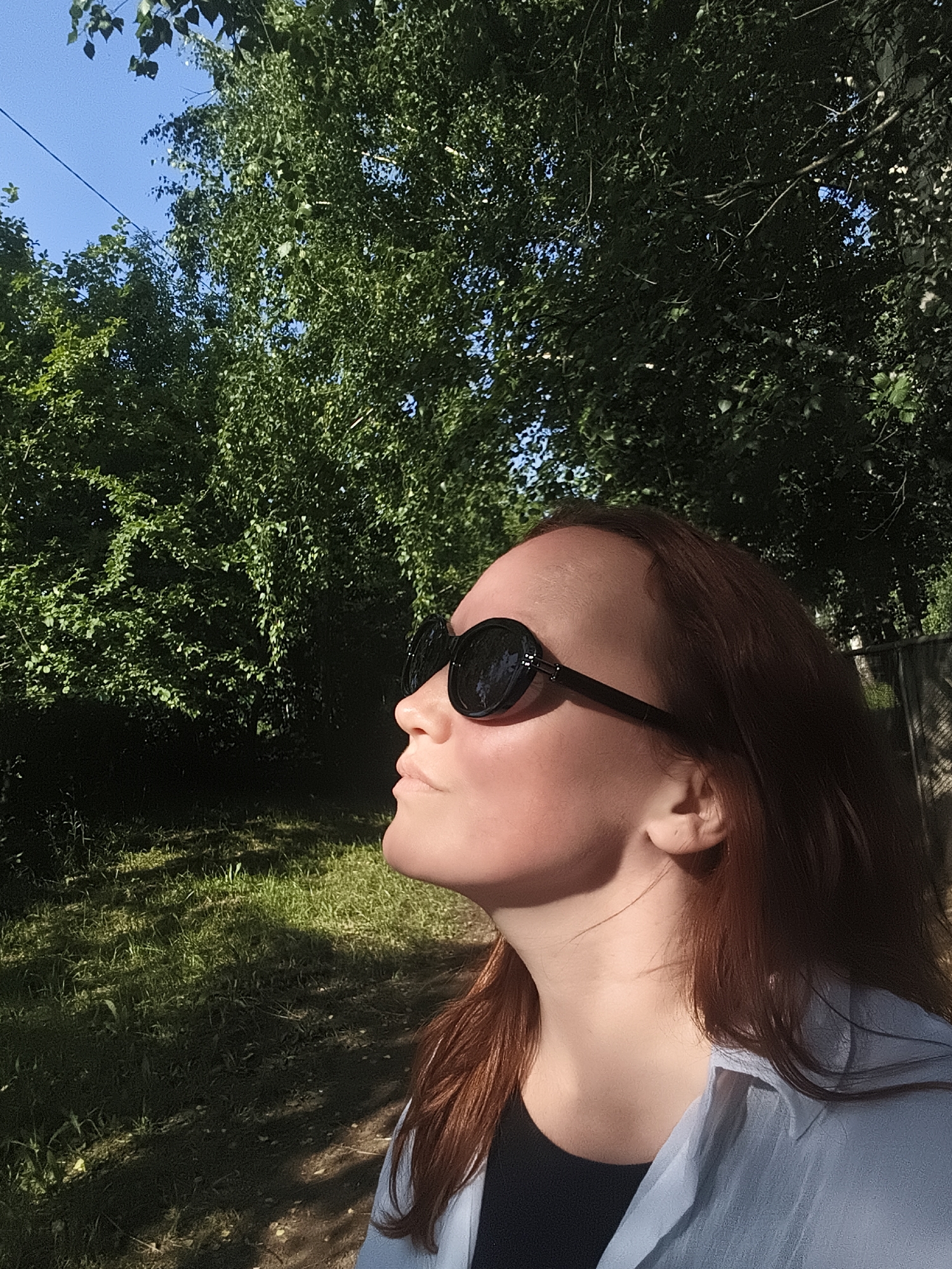 The image shows a young woman with reddish-brown hair, wearing large, dark sunglasses and a light blue shirt over a black top. She is positioned in the foreground, looking upwards and to the left, with her face illuminated by sunlight. Behind her, there is lush green foliage from trees, and a patch of sunlit grass. The setting appears to be an outdoor area, possibly a park or a garden, with a glimpse of a fence in the background. There are no discernible elements that would specifically place the image in Chaikovsky, Russia.