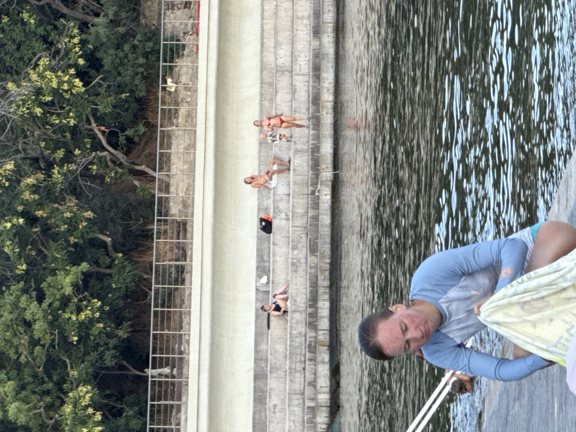 In the city of Ut'os, Ukraine, a photograph captures a scene at a waterfront with a tiered, stone seating area. In the foreground, a woman wearing a blue long-sleeved shirt and turquoise shorts crouches down, holding a light-colored towel. She appears to be drying herself or preparing to wrap up. Behind her, the rippling surface of the water is visible. Further back, on the stone steps, a group of people are present. A man in white shorts is seated on the steps, talking on a phone. A small child stands next to him. A woman in a red bikini is walking down the steps, and another woman in a black swimsuit is seated on the steps, leaning back. Bags are placed on the steps near some of the individuals. Above the seating area, a white wall and a metal railing are visible, with lush green trees and foliage forming a dense background.