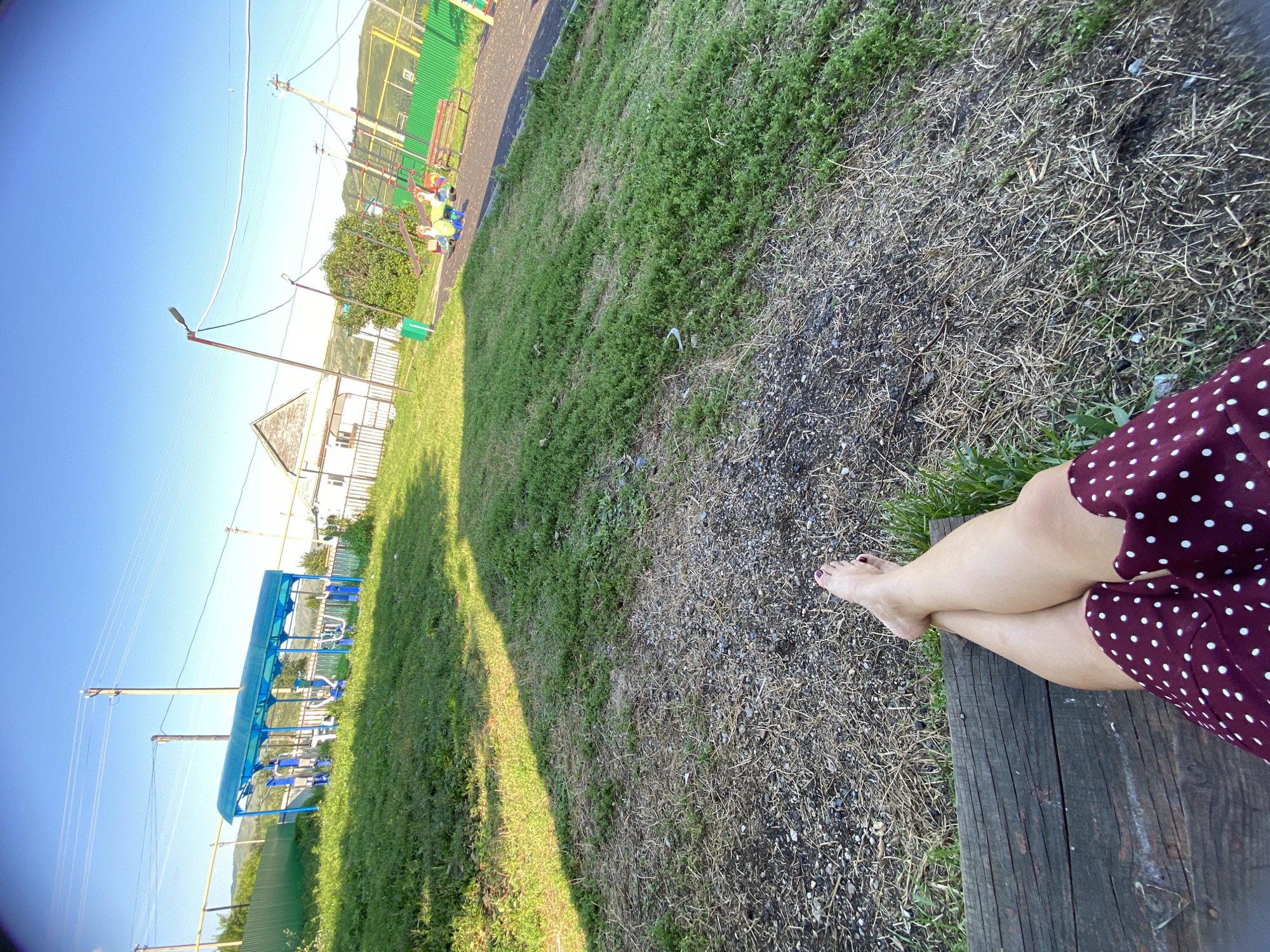 The image depicts an outdoor scene in Maloarslangulovo, Russia, likely a park or recreational area. In the foreground, a person's bare legs and feet are visible as they sit on a wooden bench. They are wearing a dark red dress with white polka dots. The ground in front of the bench is covered with gravel and dry grass.
In the middle ground, there is a grassy slope leading up to a children's playground. The playground features a blue canopy covering exercise equipment, including what appear to be weight machines. Further to the right, there are swings and other playground structures with colorful elements. A red bench is also present near the playground.
The background reveals a residential house with a light-colored facade and a dark roof, situated behind a white fence. Power lines crisscross the sky, supported by utility poles. The overall environment suggests a sunny day with clear blue skies. Rolling hills are visible in the far distance.