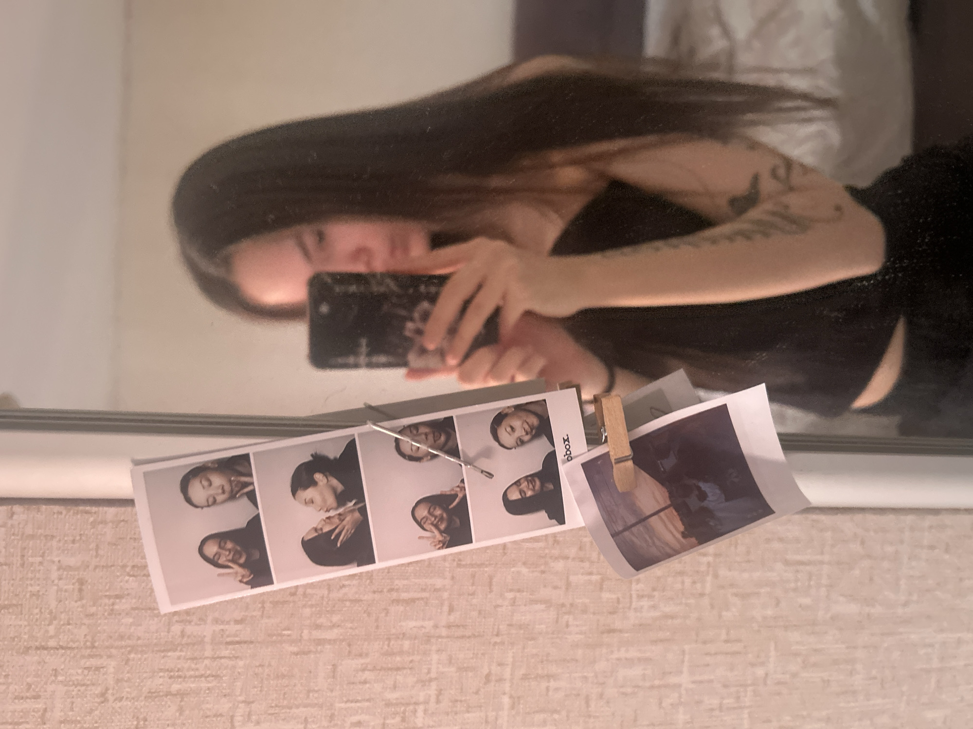 A person with long dark hair is taking a mirror selfie, holding a phone with a dark floral patterned case. They are wearing a black top and have tattoos on their left arm. The mirror is mounted on a wall with a textured, light beige wallpaper. Below the mirror is a photo booth strip of four images of the same person in various poses, attached to the wall with a silver clip. A separate photograph is also attached to the wall using a wooden clothespin. The location is possibly Chaikovsky, Russia, based on the user's query.