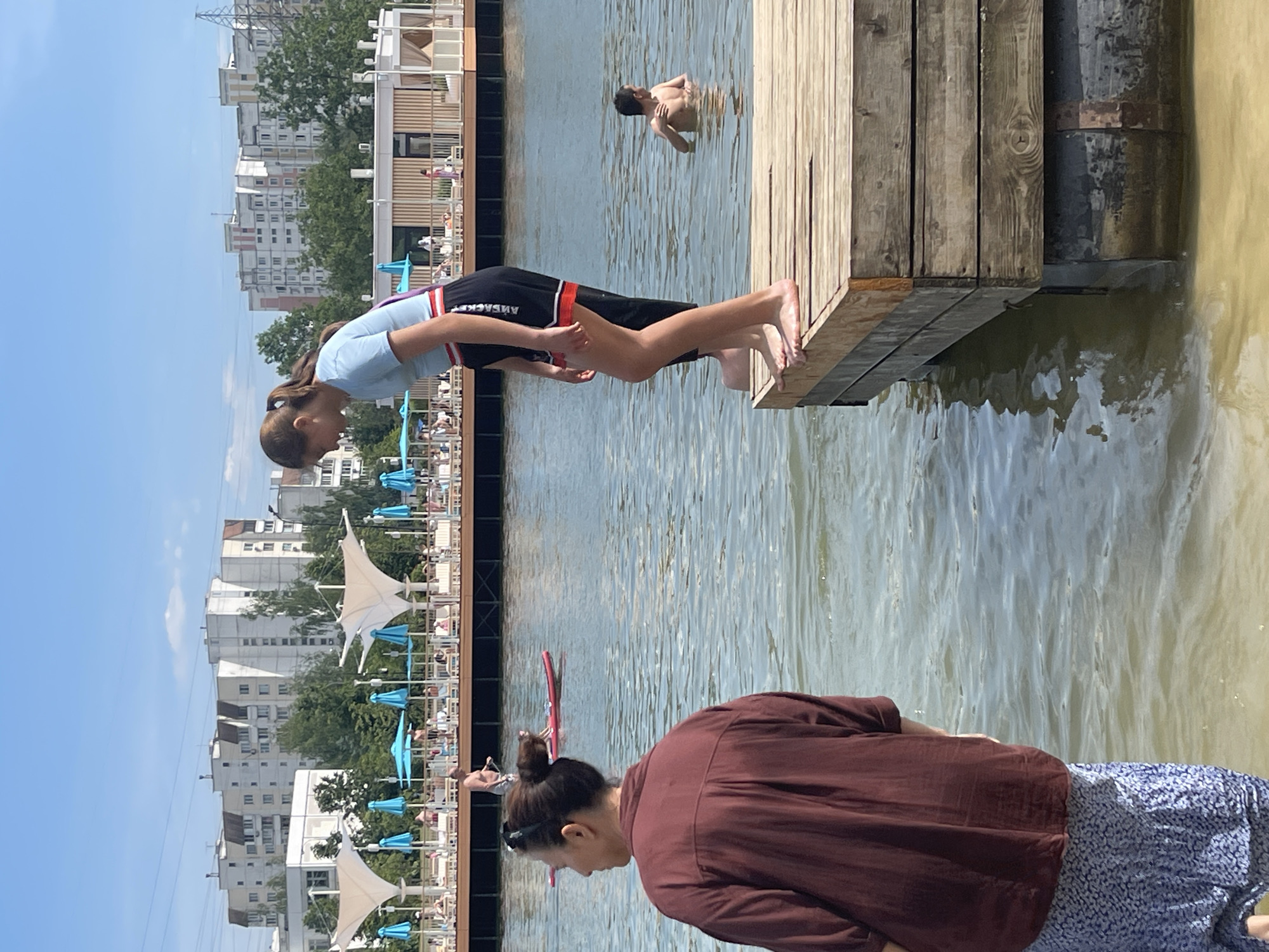 This image captures a daytime scene at Serebryany Bor Beach 3 in Moscow, Russia. The environment is an urban recreational waterfront under a clear, bright blue sky.
In the immediate foreground, to the left, a person, presumed female, is seen from behind, facing the water. She wears a long-sleeved maroon top and a blue and white patterned skirt. Her dark hair is styled in a bun. To the right, a wooden dock or floating platform extends into the water. On its edge, a young person, presumed female, stands barefoot, bending slightly forward and looking down at the water. She is dressed in a light blue short-sleeved shirt and dark shorts with red and white trim. Behind her, a second pair of bare feet is visible on the dock, suggesting another person is standing directly behind her.
In the midground, the water surface is calm, with areas of light brown closer to the shore transitioning to darker blue further out. To the right of the dock, a person, possibly a male, is partially submerged in the water, facing away from the viewer, appearing to swim or wade. Further left in the water, a man is standing on a red paddleboard, holding a paddle.
The background features an extensive recreational area across the water. This area comprises a long wooden boardwalk or platform lined with numerous white sun loungers, many occupied by individuals relaxing. Light blue sun umbrellas are interspersed among the loungers, along with several distinctive large white tent-like canopies with angular, sail-like forms. A modern building with light wooden paneling is also visible within this recreational zone. Beyond the recreational facilities, a dense line of green trees separates the area from several multi-story residential apartment buildings. These buildings exhibit typical Soviet-era or post-Soviet architecture, with light-colored facades and multiple windows. Faint power lines traverse the upper portion of the sky.