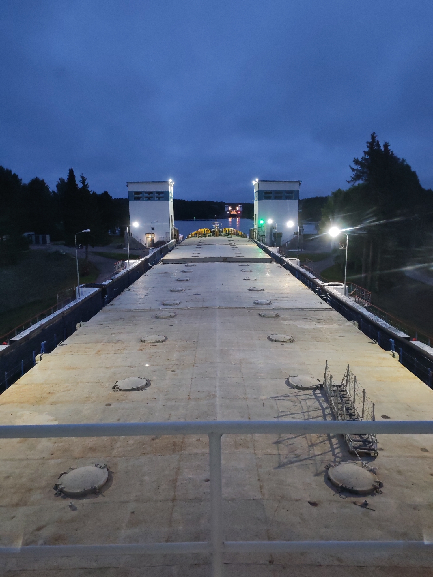 The image depicts a canal lock system at dusk, likely in or near Markovo, Russia, given the provided context. The perspective is from the deck of a large vessel, looking forward down a long, concrete-covered area. Two imposing white lock gate control towers stand prominently at the far end, illuminated by lights. A green signal light is visible on the left tower, indicating readiness or passage. The sky is a deep blue, transitioning into twilight, with a few scattered clouds.
The deck of the vessel is textured concrete with numerous circular access hatches scattered across its surface. A white metal railing runs along the foreground, partially obscuring the view of the deck. To the sides of the concrete deck, the edges of the canal lock are visible, lined with dark, textured walls. Beyond the lock structure, a line of dark trees forms the horizon, with a few more trees and some structures visible on the left bank. In the distance, on the water, another vessel with red and white lights can be seen. The overall atmosphere is serene and industrial, illuminated by artificial lights against the fading daylight.