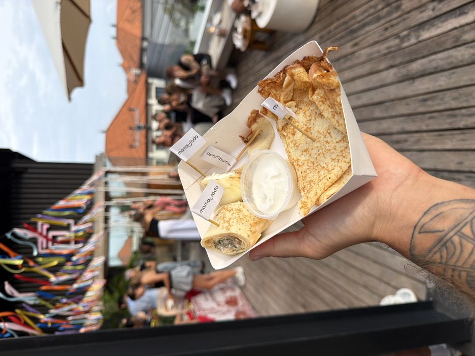 A first-person perspective image captures a hand holding a food tray at an outdoor social event on a wooden rooftop deck in Canggu, Indonesia. The hand, with a visible tattoo on its forearm, presents a white cardboard tray containing several crepes or pancakes. One crepe is rolled and filled, another section holds folded crepes, and a small clear plastic cup contains white dipping sauce. Small white flags on toothpicks, some branded "mama_soda" and one marked "3," are inserted into the food.
In the midground, a gathering of approximately 10-15 people are casually socializing. Some individuals stand near a table with a patterned cloth on the left, while others are seated on a low wooden bench or ledge on the right. Overhead, colorful ribbons are strung as decoration. A dark, slatted building structure is partially visible on the far left. The background reveals the distinctive terracotta tiled rooftops characteristic of buildings in Canggu, set against an overcast sky. A light-colored outdoor umbrella is visible on the far right. The overall scene depicts a relaxed daytime event.