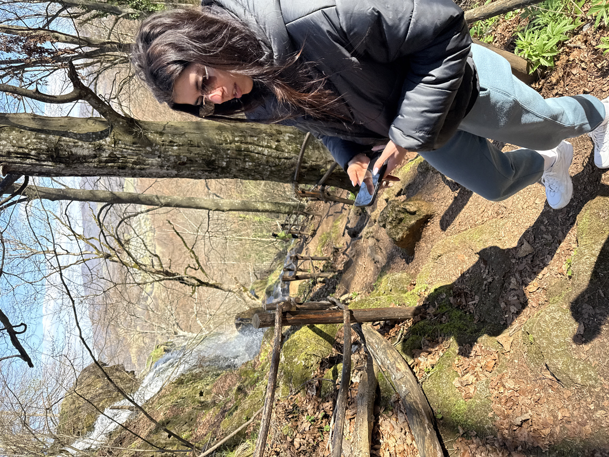 A woman in a black puffer jacket, blue sweatpants, and white sneakers stands on a rocky, leaf-covered terrain. She is holding a smartphone. Below her, a wooden ladder structure leads down to a waterfall. Trees with bare branches are visible. The location is Ribaritsa, Bulgaria.