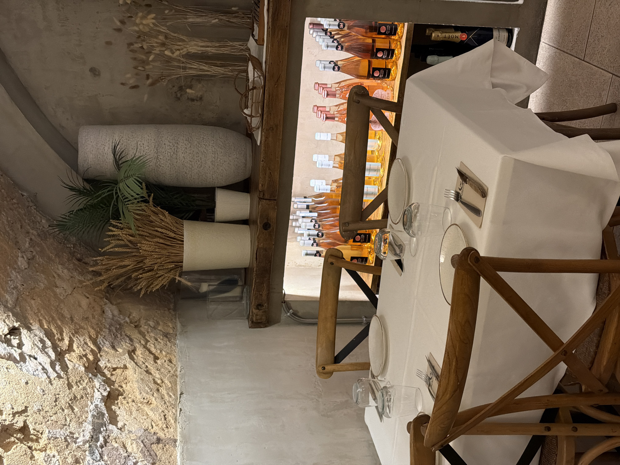 The image depicts a table setting in what appears to be a restaurant with an exposed stone ceiling and textured walls. A white tablecloth covers a table, set with plates, glasses, and cutlery. Two wooden chairs are positioned at the table. In the background, a shelf displays numerous bottles of rosé wine and a bottle of champagne. The overall ambiance is dimly lit, with a focus on the table setting and the wine display. The location is identified as Palmanova, Spain.