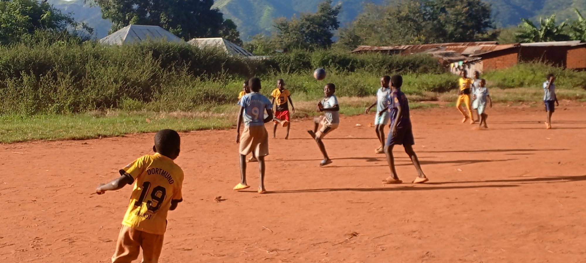 The image shows a group of children playing soccer on a red dirt field in Ngalula, Democratic Republic of the Congo. In the foreground, a child wearing a yellow Dortmund soccer jersey with the number 19 is facing away from the camera, arm extended. Several other children are in the middle ground, actively playing soccer; one is kicking a ball in the air. They are wearing various t-shirts and shorts. In the background, there are green bushes, trees, and simple buildings with corrugated metal roofs. Mountains are visible in the distance.