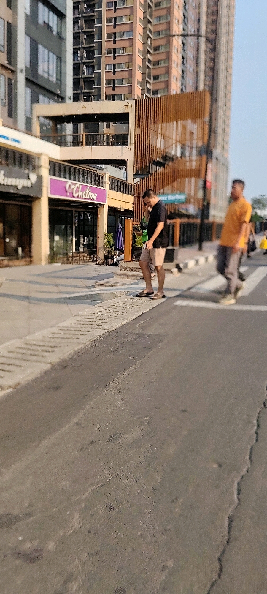 The image depicts a street scene in Jayamukti, Indonesia, with a focus on pedestrians and commercial buildings. In the foreground, a man wearing a black t-shirt and light-colored shorts stands on a crosswalk, looking at his phone and talking on it. He is wearing flip-flops. To his right, another man in an orange t-shirt is walking across the same crosswalk. In the background, there are several modern buildings, including one with a patterned wooden facade. A sign for "Chatime" is visible on one of the lower floors of a building. The sky is clear and blue, suggesting it is daytime. The street appears to be paved and well-maintained, with some cracks and imperfections visible. There is a concrete curb and a patterned walkway or sidewalk area next to the road.