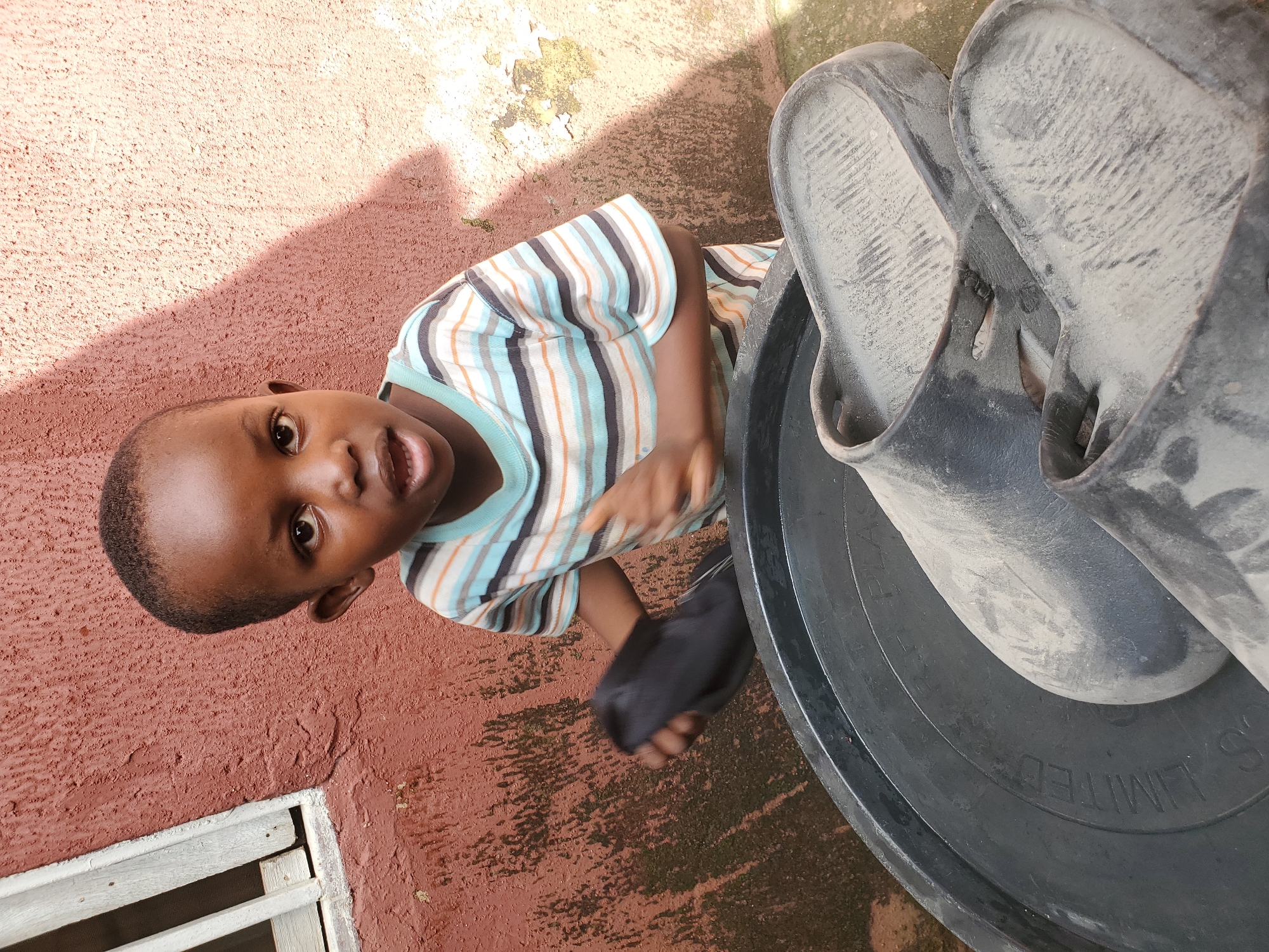 A young Black child, likely a boy, is the central subject of this image, which appears to be taken outdoors. He is wearing a horizontally striped t-shirt with colors of white, brown, light blue, and orange. The child is looking up and towards the camera with an open mouth and a curious expression.
Beside the child is a large, dark-colored basin or tub, and inside it are a pair of dusty black flip-flops. The ground appears to be made of concrete or a similar hard surface, with a reddish-brown wall behind the child. The wall has a textured finish and shows some signs of wear and discoloration. Sunlight is casting a shadow on the wall, suggesting it is daytime, likely mid-morning or afternoon. A portion of a wooden-slatted window frame is visible in the lower left corner of the image.
The overall impression is one of a casual, everyday moment, possibly in a residential area in Jos, Nigeria, given the provided context. The child's expression is engaging and full of life. The presence of the flip-flops and the dusty basin hint at outdoor activities or perhaps chores. No visible text is discernible in the image.