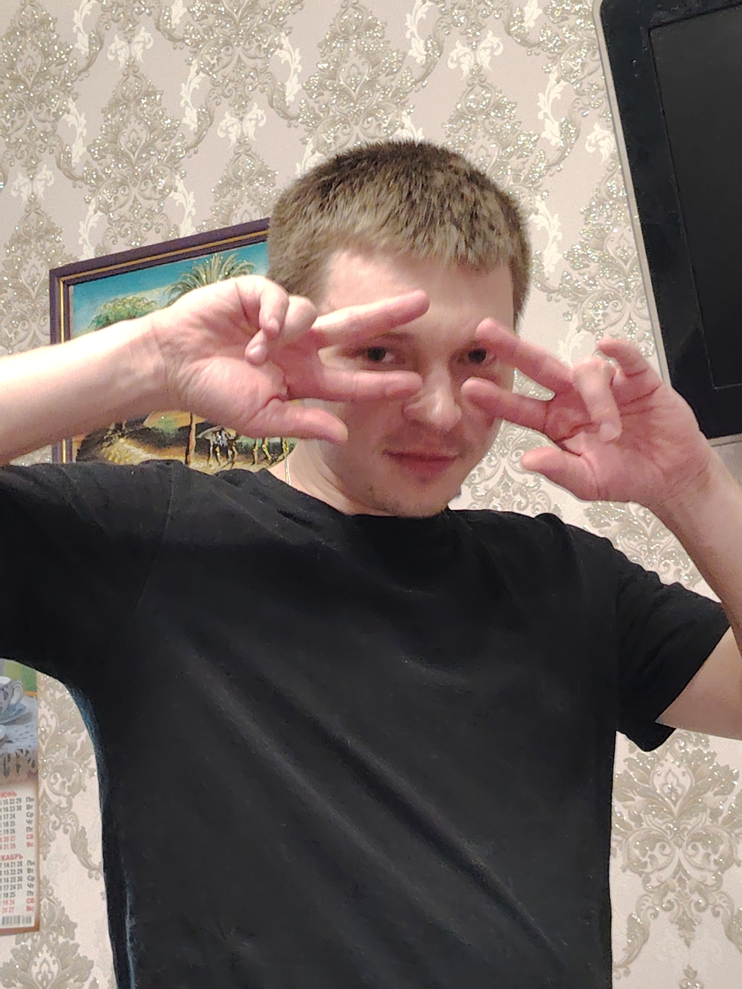 A fair-skinned person, likely male, poses for a selfie indoors, facing the camera directly. They are wearing a black t-shirt and making a "peace" or "victory" sign with both hands, positioning the signs near their eyes. The person has short, light-brown hair.
The background is a room with patterned wallpaper in light beige and silver. A framed picture of a landscape is partially visible behind the person's left hand. A dark-colored, rectangular object (possibly a television) is seen on the upper right side of the frame. To the left, partially visible, is a wall calendar. Based on the Cyrillic text, the months June and October are visible.
The OCR extracted the following text from the calendar:
SHO
12229
16 23 30
1724
1825
1925
2027
КАБРЬ
14 21 28
15 22 29
142330
17 24 31
1825
The presence of the Cyrillic alphabet and the reference to months like "КАБРЬ" (October) suggest the photo was taken in a Russian-speaking region.
Based on visual evidence, there is no definitive indication this photograph was taken in Noviy, Russia.