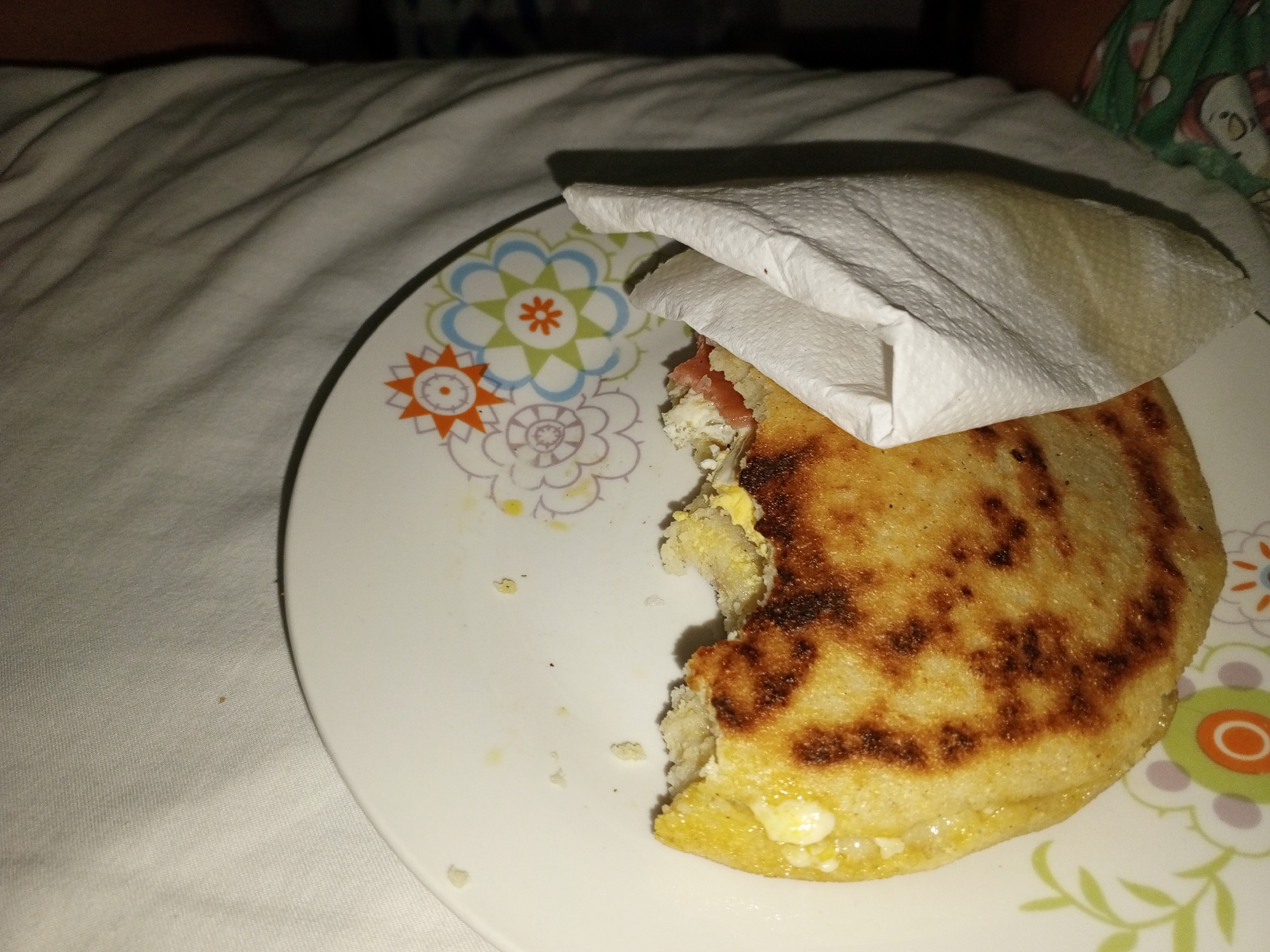 The image captures a close-up view of a partially eaten meal resting on a white, crumpled bedsheet or blanket. Dominating the center is a round, white plate adorned with a floral pattern featuring orange, green, blue, and purple flowers. On the plate sits a golden-brown arepa, a traditional corn cake, with a significant bite taken out of its left side. The arepa's surface shows grill marks, indicating it was cooked on a griddle.
The exposed filling of the arepa reveals white, crumbly cheese and hints of a reddish ingredient, possibly ham or tomato. A white, crumpled paper napkin is placed on top of the arepa, partially covering the filling and showing a yellowish stain.
In the dimly lit background, the textured white fabric of what appears to be a bedsheet or blanket is visible. To the far right, a sliver of a person is discernible, wearing a garment with a green and white pattern, suggesting an indoor setting, likely a bedroom. The overall dim lighting indicates that the photo was taken at night or in a poorly lit environment. The presence of an arepa strongly suggests a Latin American context, consistent with the provided location of Porlamar, Venezuela. No text is visible in the image.