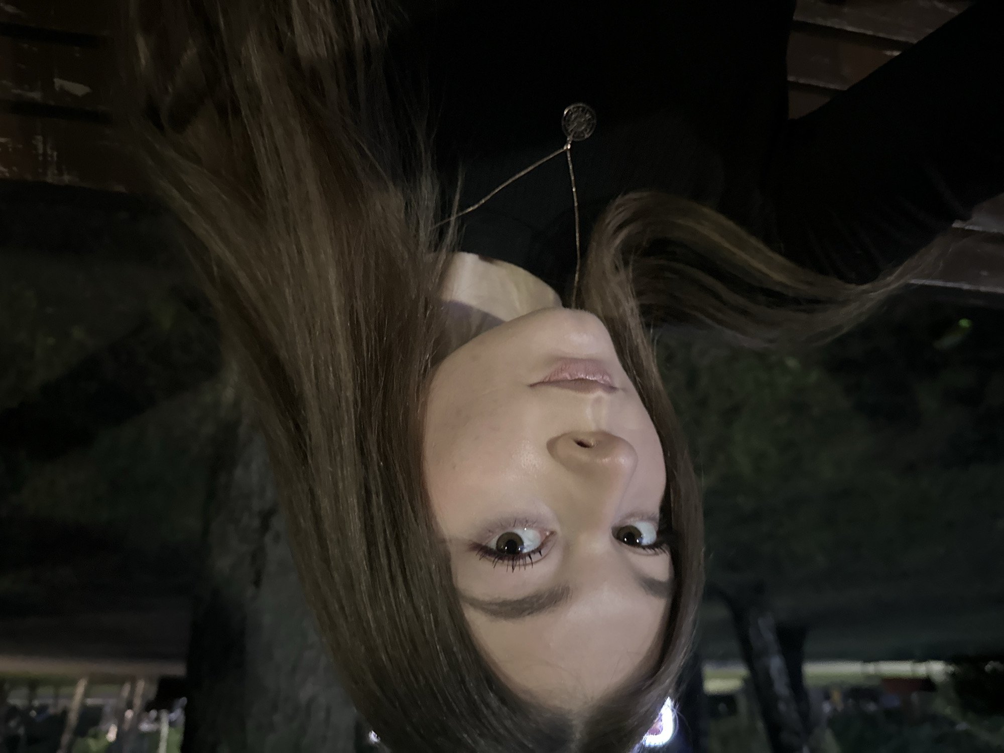 This media file is a rotated portrait-style photograph of a young woman, appearing to be taken at night. The image is rotated 90 degrees counter-clockwise, making the woman's head appear on the right side of the frame, looking towards the top-left corner.
The subject is a woman with long, straight, medium-brown hair that frames her face. She has light skin, large brown eyes, and appears to be wearing light makeup, including mascara and a subtle lip color. Her eyes are wide open and directed straight at the camera, conveying a sense of surprise, curiosity, or playfulness. She is wearing a dark, possibly black, top and a delicate gold-colored necklace with a small, circular, intricate pendant.
The scene is dark and appears to be outdoors or in a dimly lit indoor space. The background is mostly out of focus, showing blurred shapes of what seem to be trees or foliage, and some indistinct structures or lights, consistent with a night-time urban or park setting. There's a dark, possibly wooden, structure visible above her head (due to the rotation), which could be the underside of a bench or an overhead beam.
The photograph captures a direct gaze, suggesting a selfie or a close-up portrait taken by someone directly in front of her. The time of day is clearly night due to the low light conditions and the focus on the subject's illuminated face against the dark background. No visible text is present in the image. The location context provided is Kazan, Russia, though no specific landmarks are discernible.