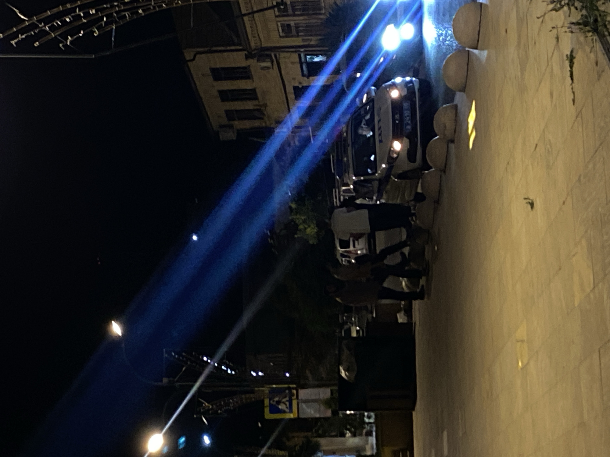 The image shows a night scene on a city street, possibly in Derbent, Russia, indicated by the presence of a police car. A white police car with its headlights and possibly emergency lights on is parked on the right side of the frame. There are several people standing or walking near the car, though their faces are not clearly visible. The street is paved with large square tiles. In the background, there is a building with windows and lights. Several streetlights are visible, casting beams of blue and white light across the scene, creating streaks and highlights. There are also some rounded concrete bollards lining the street. The overall impression is of an active nighttime scene with a police presence.