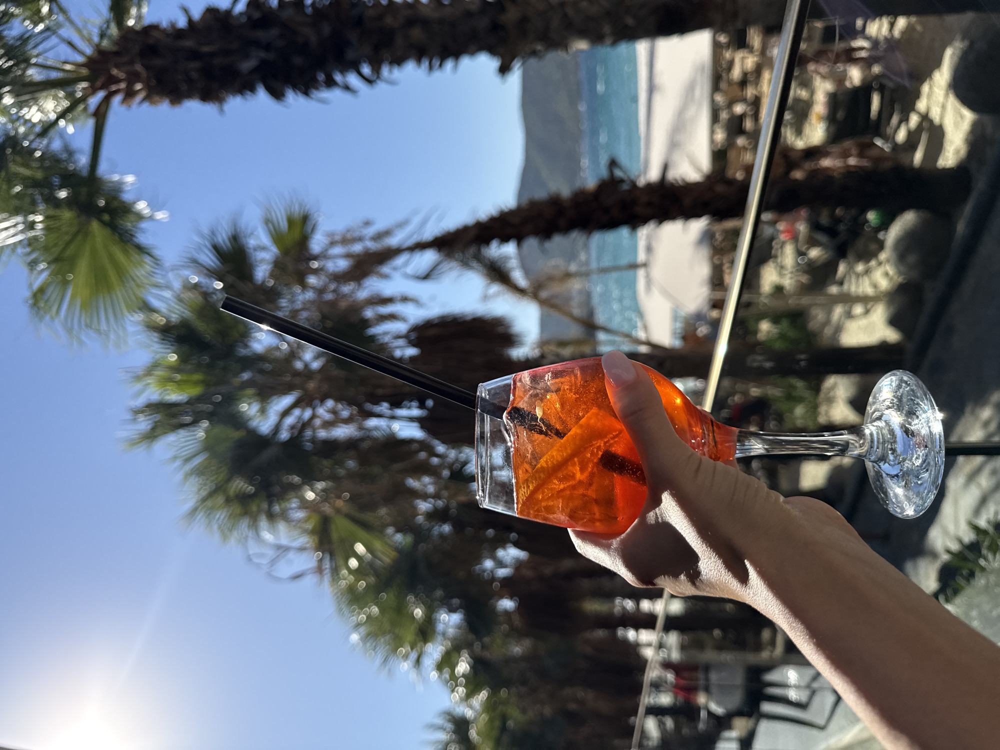 A person's hand holds a stemmed glass filled with an orange-colored beverage and a black straw. The glass contains ice and orange slices. The background features a blurred scene of palm trees, a beach, and the sea under a clear blue sky with bright sunlight. The location is identified as Bodrum, Turkey.