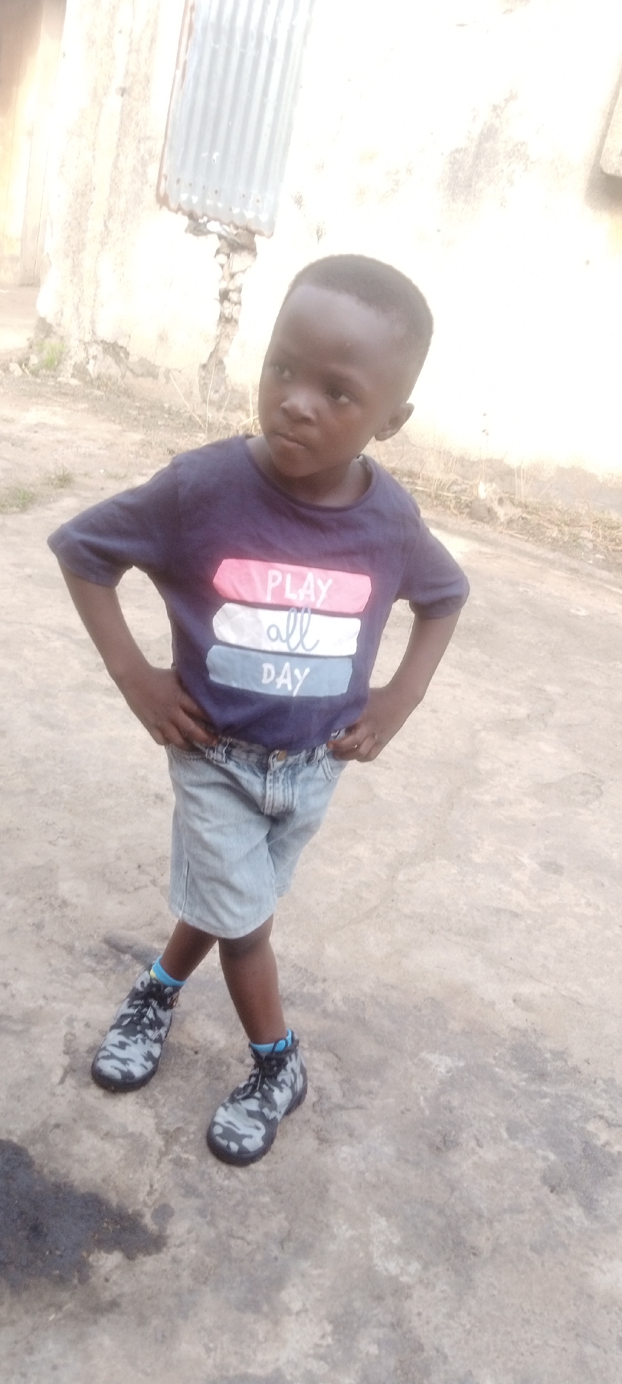 A young boy stands outdoors. He is wearing a blue t-shirt with the words "Play All Day" in white, pink, and blue stripes, light denim shorts, and camouflage sneakers with blue socks visible. His hands are on his hips. The ground appears to be concrete, with some dark staining. Behind him is a building or wall with a piece of corrugated metal affixed to it. The location is Sabon Tasha, Nigeria.
