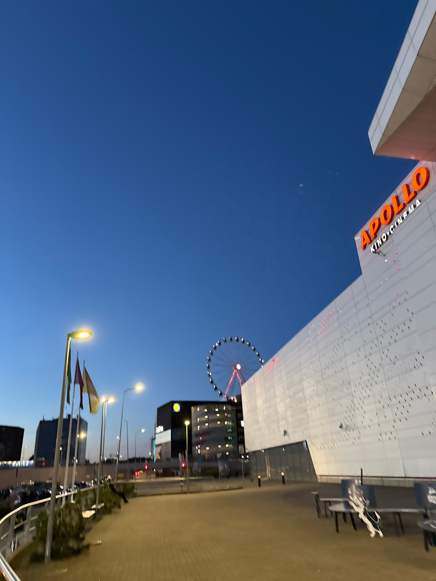 The image depicts an exterior view of the Apollo Kino cinema at the Ülemiste Keskus shopping mall in Tallinn, Estonia, during the evening. The bright orange "APOLLO KINO CINEMA" signage is prominently displayed on the modern white facade of the building. A large Ferris wheel, lit with red lights, is visible in the background to the left. The sky is a deep blue, indicating dusk. In the foreground, a paved walkway leads towards the cinema. Streetlights illuminate the area, and several flags are mounted on poles. Other buildings, including a dark structure with yellow lights and a building with a yellow circle logo (likely a Lidl supermarket), are visible in the mid-ground. Parked cars are present in a lot to the left. There are no individuals clearly identifiable in the image, though a faint silhouette of a person can be seen on a bench in the lower right quadrant.