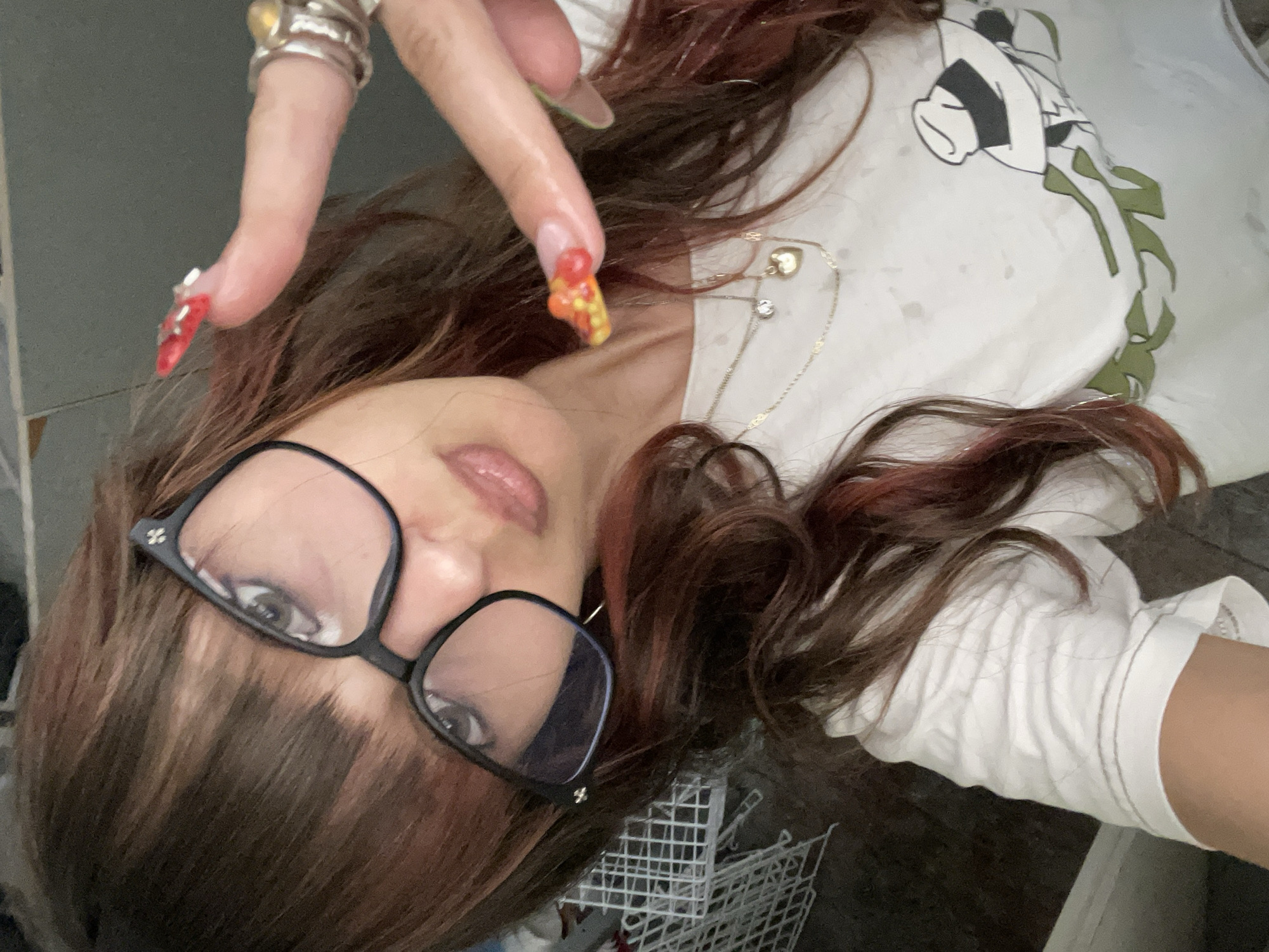 A woman with long, dark, wavy hair, some strands of which have reddish highlights, is taking a selfie. She is wearing large, black-rimmed glasses with a small silver embellishment on the top corner of the left lens. Her eyes are visible through the lenses, and she has on noticeable makeup, including lipstick and possibly eyeshadow.
She is wearing a white t-shirt with a graphic design on it. The design appears to be a cartoon character wearing a hat, with green elements that could be leaves or branches. She is also wearing two necklaces, one with a small clear stone pendant and another with a gold heart pendant.
Her right hand is raised, and she is gesturing with two fingers in a peace sign. Her fingernails are long, elaborately decorated with red, yellow, and white patterns, resembling a strawberry design. She is also wearing several silver rings on her fingers.
The background is out of focus, but appears to be an indoor setting. To the left, there is a white wire rack, possibly a laundry basket or storage unit. The wall behind her is a neutral color.
The image does not contain any information that would allow for identification of the location within the city of Raanana, Israel.
