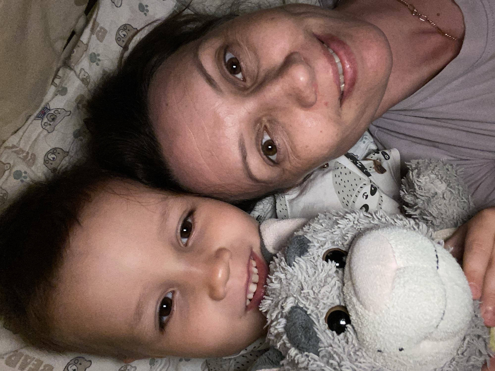 A woman and a young child are lying on a bed, smiling at the camera. A plush toy is also visible, positioned close to the child. The bed has a patterned sheet featuring cartoon animal motifs. The location, Chaikovsky, Russia is not visible in the picture.