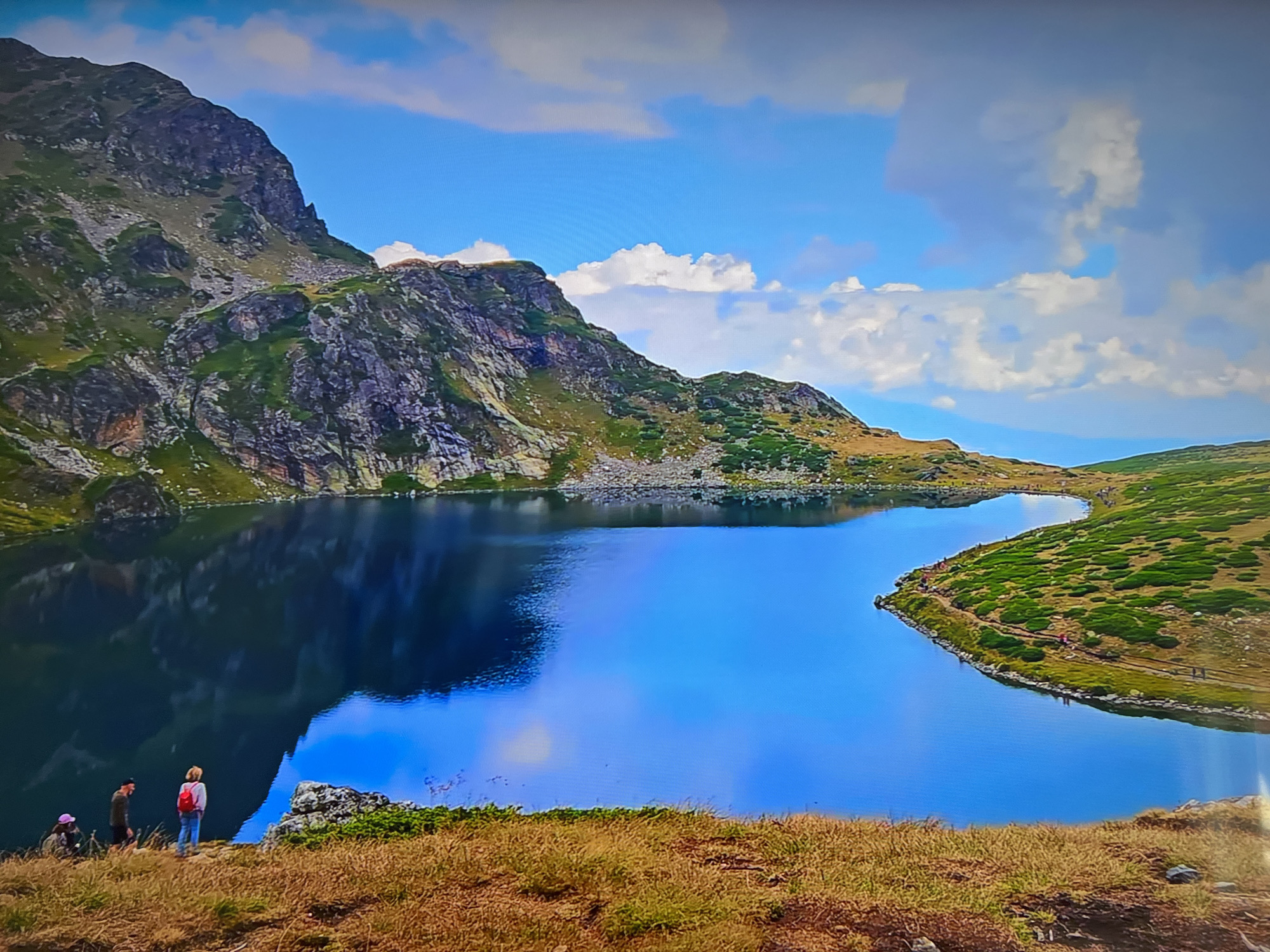 The image shows a mountain lake surrounded by rugged terrain. A group of three people stand on a grassy bank overlooking the water. The lake reflects the sky and surrounding mountains. The landscape consists of mountains, grass, and a partly cloudy sky. There is a visible trail running along the shoreline on the right side of the frame. The location is Unknown, Unknown.