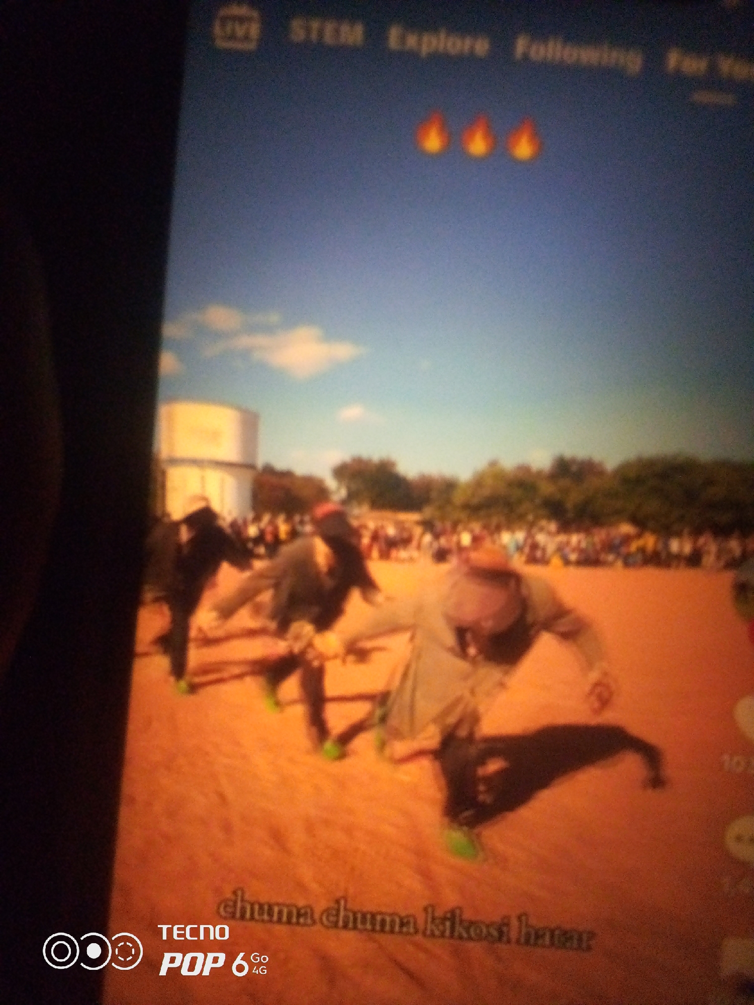 The image displays a screen recording of a social media video. In the foreground, three individuals are dancing or performing a coordinated movement in what appears to be an outdoor setting. The ground is covered in a reddish-brown dirt. The dancers are wearing dark clothing, and one person in the center is wearing a hat. In the background, a crowd of people is gathered, and a large, cylindrical structure, possibly a water tower, is visible. The sky is blue with scattered clouds. The bottom of the screen shows branding for "TECNO POP 6 4G" and text that appears to read "chuma chuma kikosi hatar". The top of the screen includes UI elements like "LIVE", "STEM", "Explore", and "Following". The overall impression is that of a performance or event captured on a mobile device. The location of Howayi, Nigeria is not discernible from the visual information present in the image.