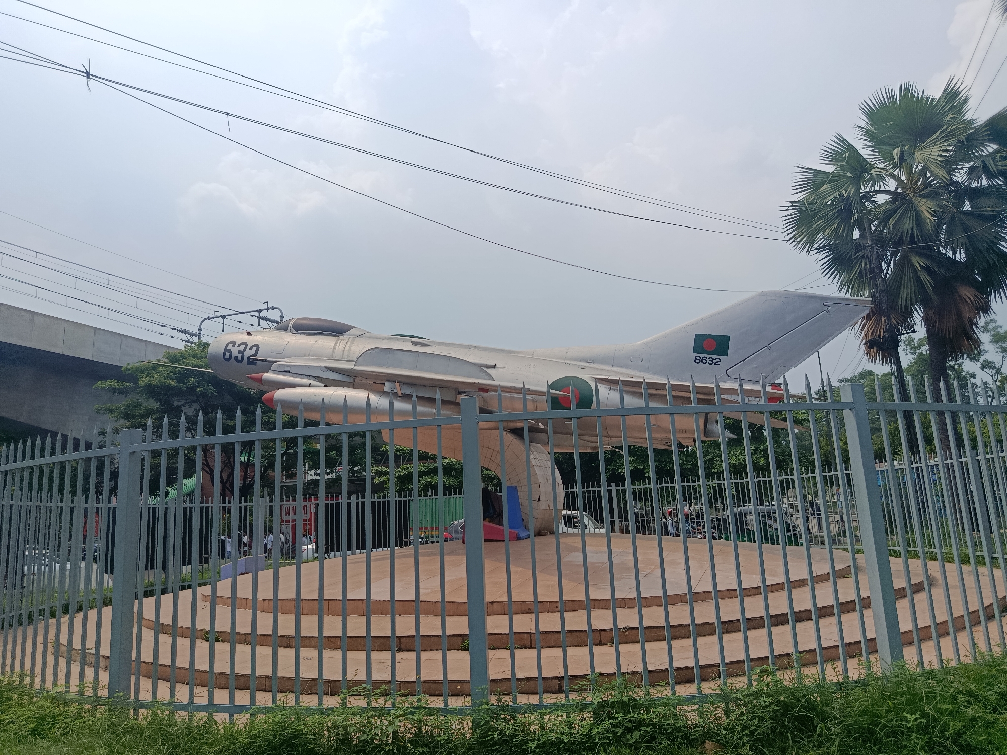 A silver Mikoyan-Gurevich MiG-21 fighter jet is on display, mounted on a concrete pedestal that is surrounded by a gray metal fence. The jet is marked with the number "632" on its nose and a green and red roundel on its tail, which also displays the number "8632." The pedestal has several tiers, and the jet appears to be angled upwards. In the background, there is greenery, including a palm tree, and a blurred structure resembling an overpass. Power lines stretch across the sky. The scene is captured in daylight with overcast skies. The location is likely Dhaka, Bangladesh, based on the markings on the jet. People and vehicles are visible in the background, partially obscured by the fence and surrounding vegetation.