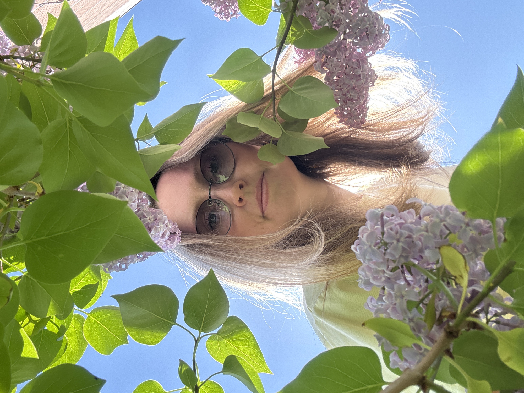 A woman with light brown hair and round glasses is looking down at the camera, framed by green leaves and lilac flowers. The clear blue sky is visible in the background. The specific location in the city of Chaikovsky, Russia is not discernible from the image.