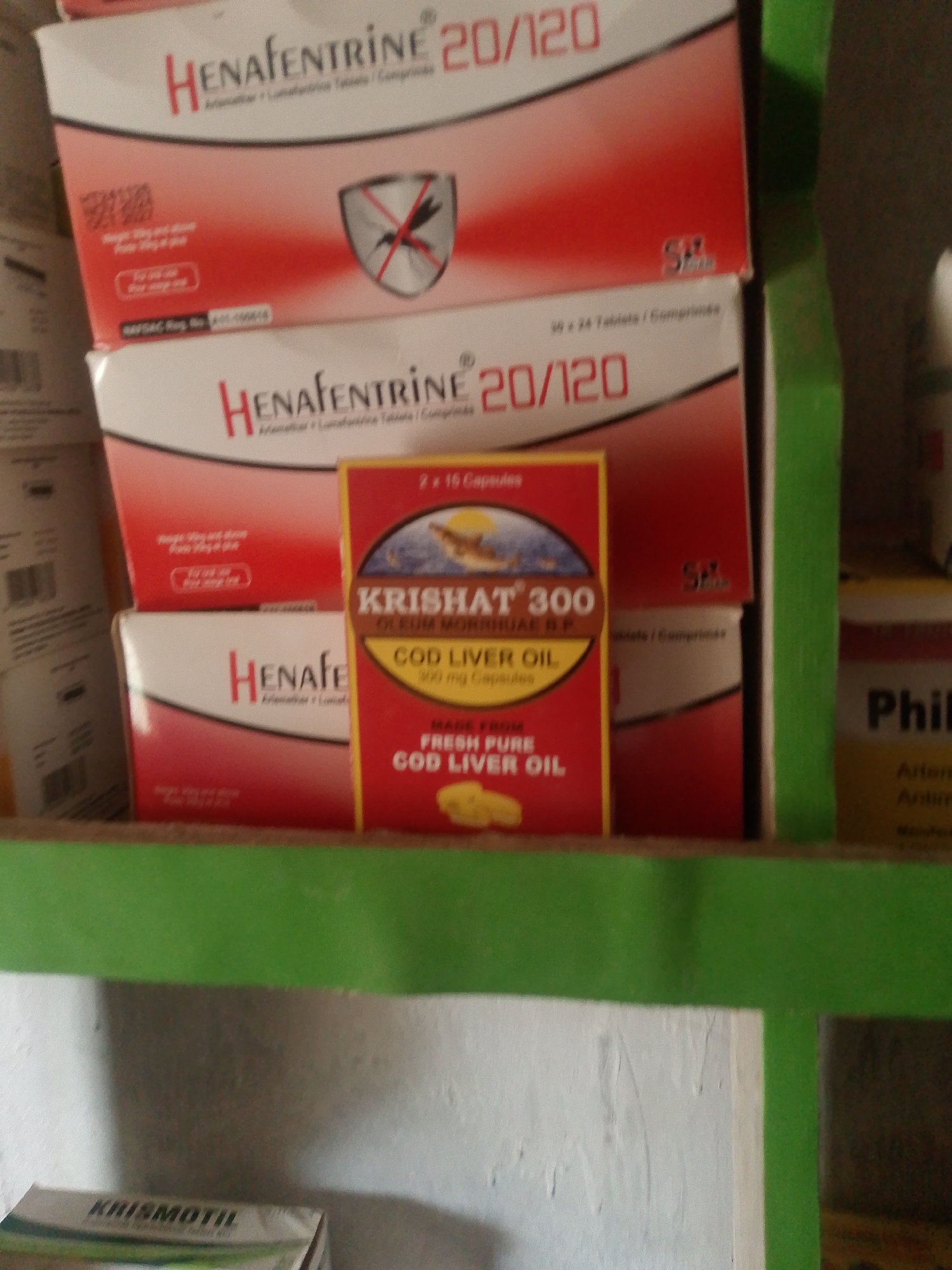 The image shows a close-up view of several boxes of medicine on a shelf. The medicine boxes are predominantly red and white, with some featuring a green accent on the shelf. In the foreground, a box labeled "KRISMOTIL" is partially visible at the bottom.
The main focus is on several boxes of "HENAFENTRINE 20/120," an antimalarial medication. One box also features a shield with a mosquito symbol crossed out, indicating its use against mosquitoes. A smaller, bright yellow and red box in the center is labeled "KRISHAT 300 OLEUM MORRHUAE B.F. COD LIVER OIL 100 mg Capsules." This box highlights the ingredients and benefits of the cod liver oil.
To the right, a box with the text "Phil" is visible, suggesting another type of medication. The setting appears to be a pharmacy or a medical store, likely in Jauro Yinu, Nigeria, as indicated by the location context. The lighting suggests an indoor environment, possibly during the day. The overall impression is one of a retail display of pharmaceutical products.