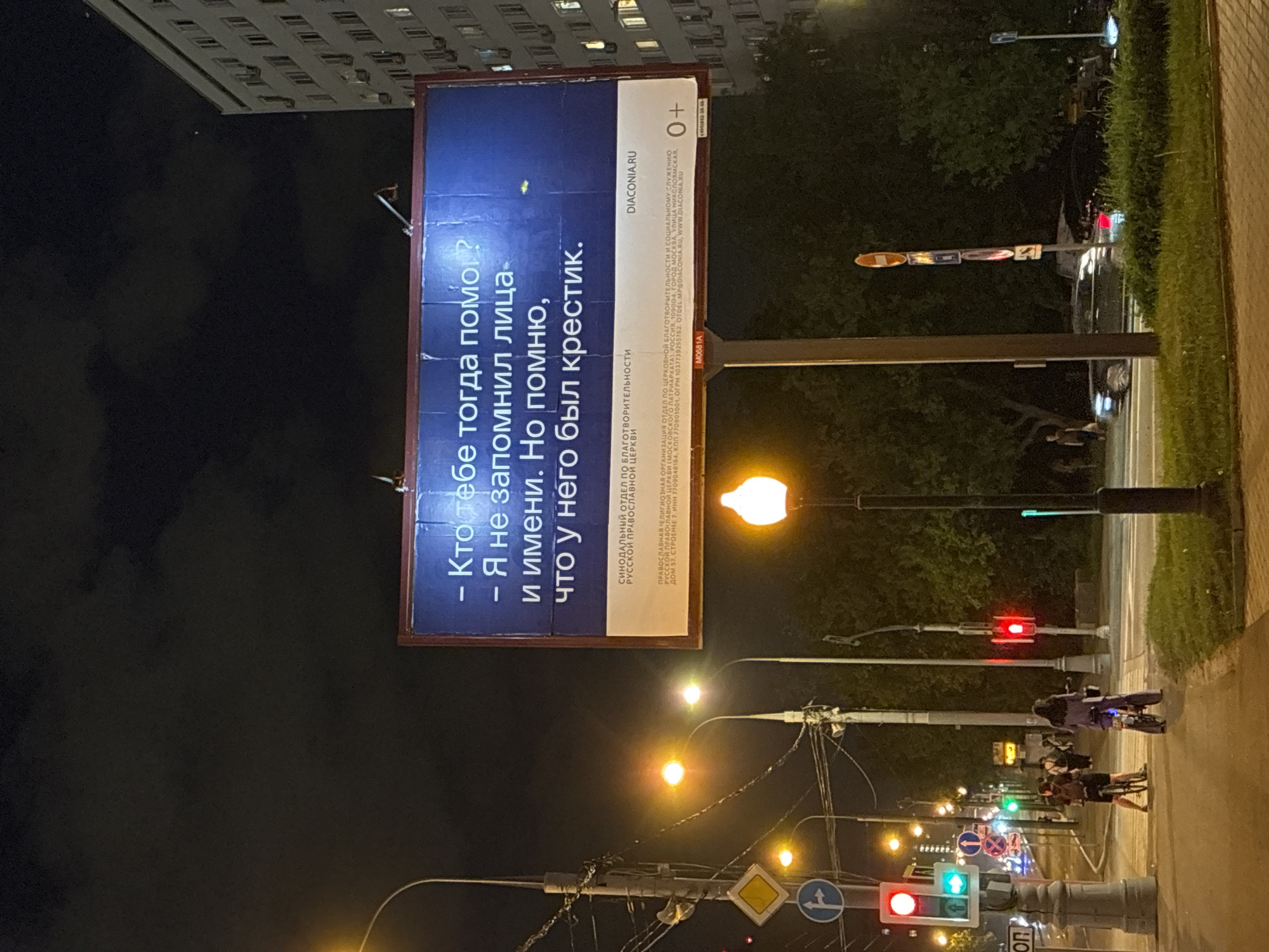 A nighttime image shows a billboard with a religious message in Russian, located in the city of Moscow, Russia. The billboard displays a question and response: "Кто тебе тогда помог? - Я не запомнил лица и имени. Но помню, что у него был крестик." Translated, this reads "Who helped you then? - I did not remember his face and name. But I remember he had a cross."
The board also contains information about the "SYNODAL DEPARTMENT FOR CHARITY OF THE RUSSIAN ORTHODOX CHURCH" and the website "DIACONIA.RU".
The street scene features a pedestrian walkway on the right, with people walking, some with bicycles. Streetlights are visible along the road, with traffic lights indicating red and green signals. Overhead electrical wires are strung across the sky. In the background, a tall building is partially visible under the night sky.