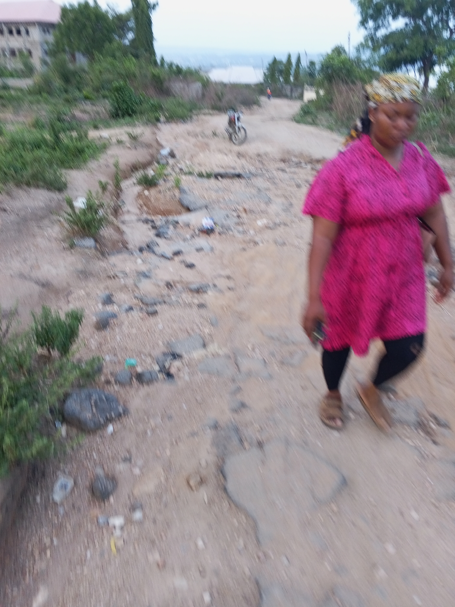The image shows a woman walking on a dirt road in Zango-ndaji, Nigeria. She is wearing a pink patterned dress, black leggings, and sandals, and a head covering. There are buildings and trees in the background. In the distance, a person can be seen riding a bicycle on the road. The road surface is uneven with rocks and debris scattered about. The lighting suggests it is daytime.