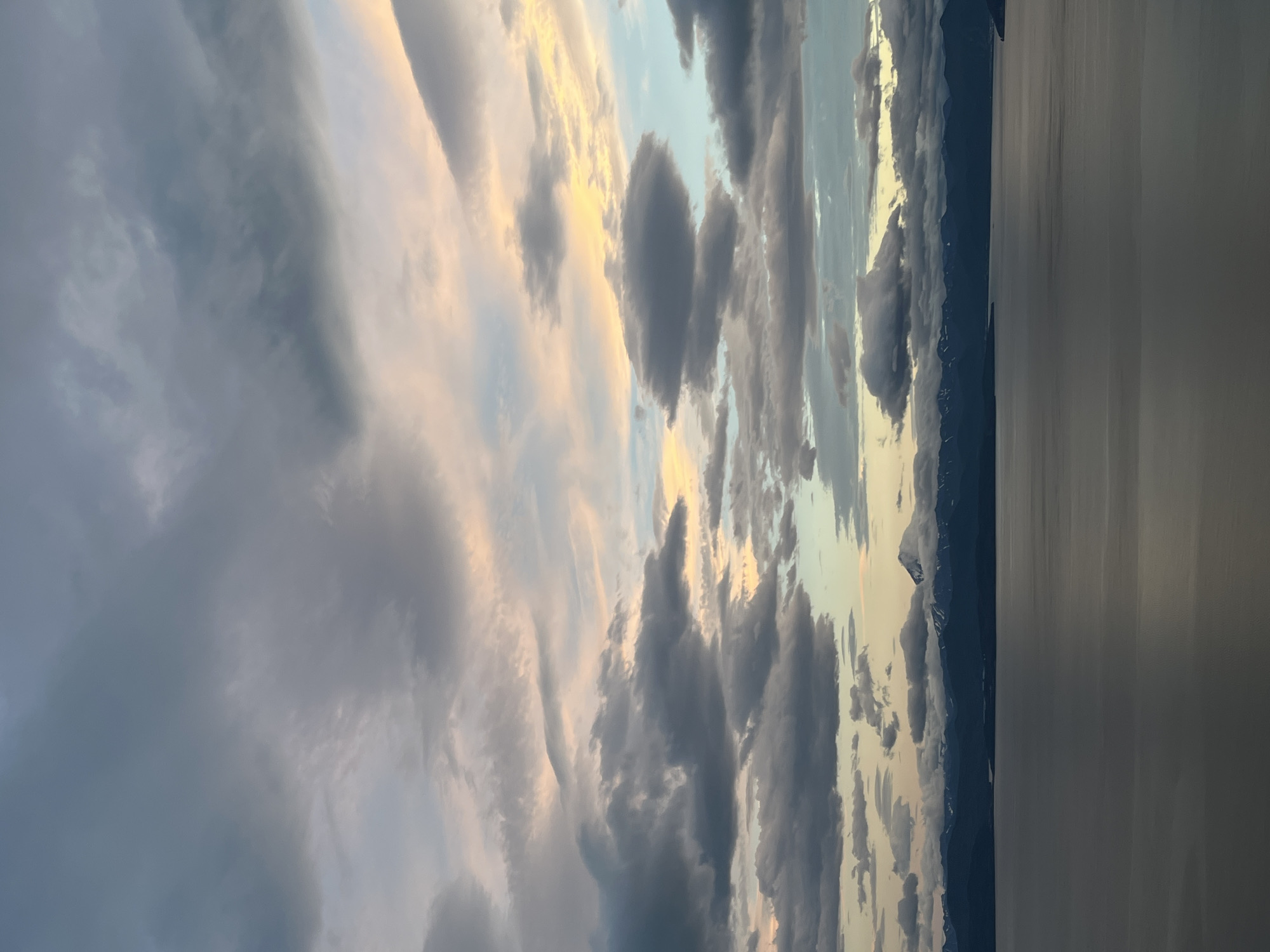 The image shows a panoramic view of a large body of water with a mountainous coastline under a dramatic sky. The water is dark and reflects the clouds and the muted light. The coastline is lined with dark, silhouetted mountains, some of which are snow-capped. A prominent, cone-shaped mountain with a snow-covered peak stands out against the backdrop of other mountains. The sky is filled with a complex arrangement of clouds. Dark, heavy clouds dominate the lower and middle sections of the sky, interspersed with lighter, wispy clouds. Towards the upper right, there are streaks of soft pink and yellow light, suggesting either sunrise or sunset. The scene evokes a sense of natural grandeur and possibly remoteness. The location of Petropavlovsk-Kamchatsky, Russia is not discernible from the image itself. There are no people, actions, or specific objects that would indicate a city setting or any particular activity.