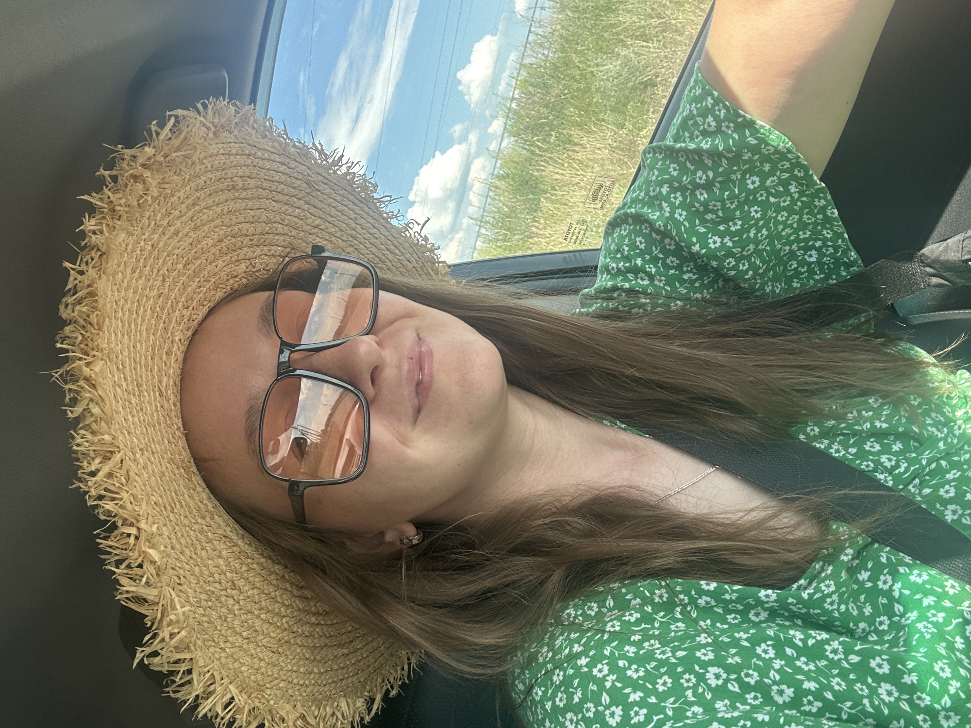 A young woman wearing a wide-brimmed straw hat and oversized sunglasses is in the driver's seat of a car, looking at the camera and smiling. She has long, brown hair and is wearing a green dress with a white floral pattern. A seatbelt is visible across her chest. The background shows a sunny sky with clouds and green fields, suggesting an outdoor setting, possibly on a road trip. The image does not contain any specific visual information to determine the location within Bely Yar, Russia.
