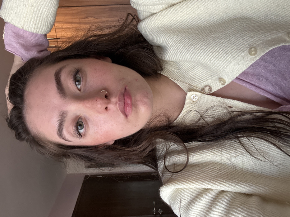 A young woman with light skin, brown hair, and green eyes looks directly at the camera. She has a nose ring and freckles. She is wearing a light purple top under a cream-colored cardigan. Her hair is down and slightly messy. One hand is visible holding her head. A glimpse of a dark door and light-colored wall is in the background, suggesting an indoor setting. The photo has warm lighting.