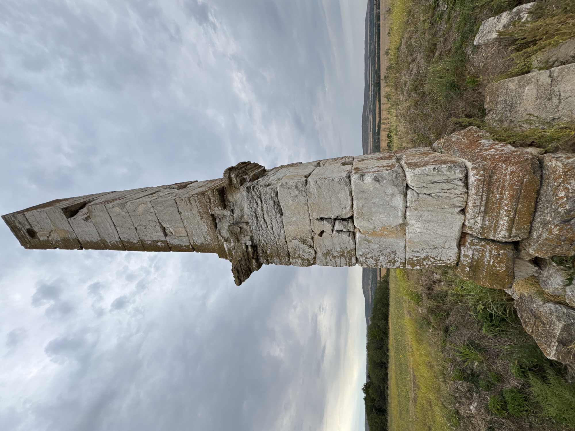 The image depicts an ancient stone monument situated in a rural landscape. The monument, tall and rectangular, is constructed from large, weathered stone blocks, showing signs of age and erosion. Its base appears to be a collection of more fragmented stones, with some exhibiting lichen growth. The monument is positioned on a slight incline, surrounded by dry grass and vegetation. In the background, there is a line of trees separating the immediate grassy area from a field of yellow flowers, possibly sunflowers. Beyond the fields, rolling hills are visible under a cloudy, overcast sky. The scene is located in the area of Lesicheri, Bulgaria. There are no people or animals present in the image.