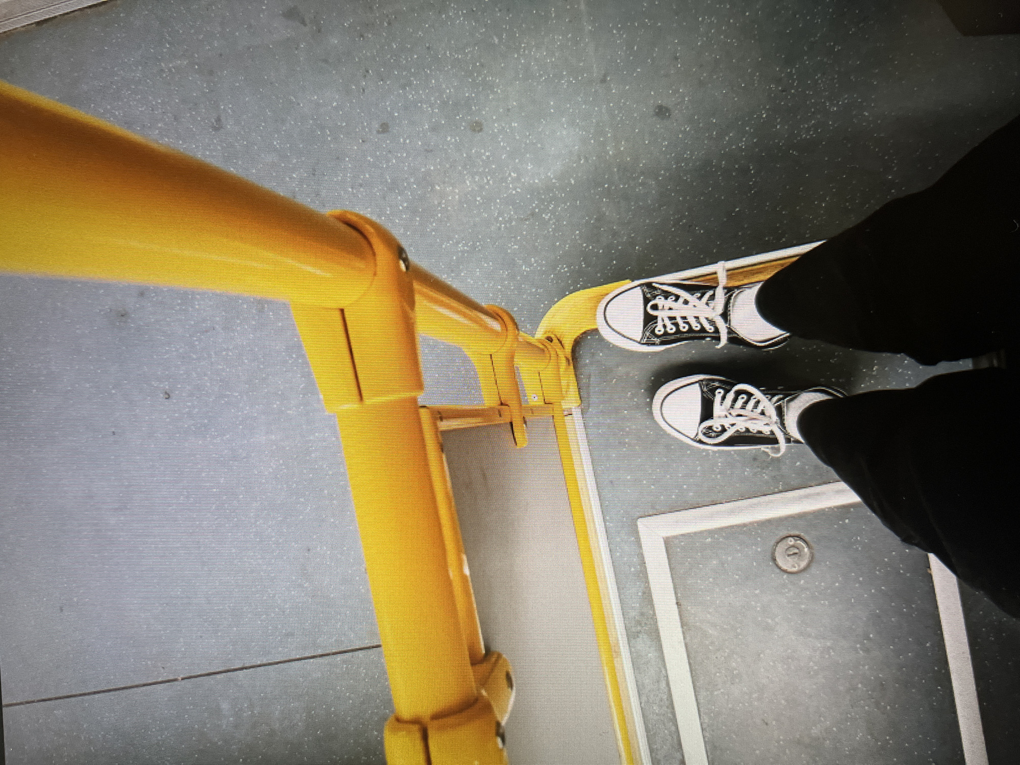 The image shows a first-person perspective of someone standing on a bus or train. The foreground includes a vertical yellow metal support pole and a horizontal yellow handrail. The floor is grey with a textured, speckled pattern. The person's legs and feet are visible wearing black pants and black and white Converse sneakers. A square access panel with a white border and a circular lock is visible in the floor. The location is consistent with public transportation in a city; however, there is no definitive evidence within the image to determine the specific city of Minsk, Belarus.
